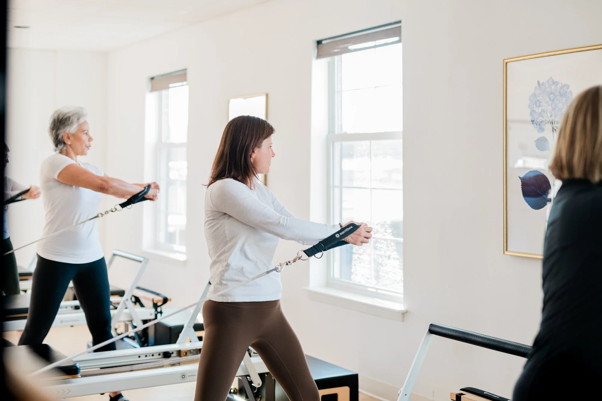 Women participating in a Pilates class at The Pilates Place in Timonium using reformer machines in a bright studio with white walls and large windows.