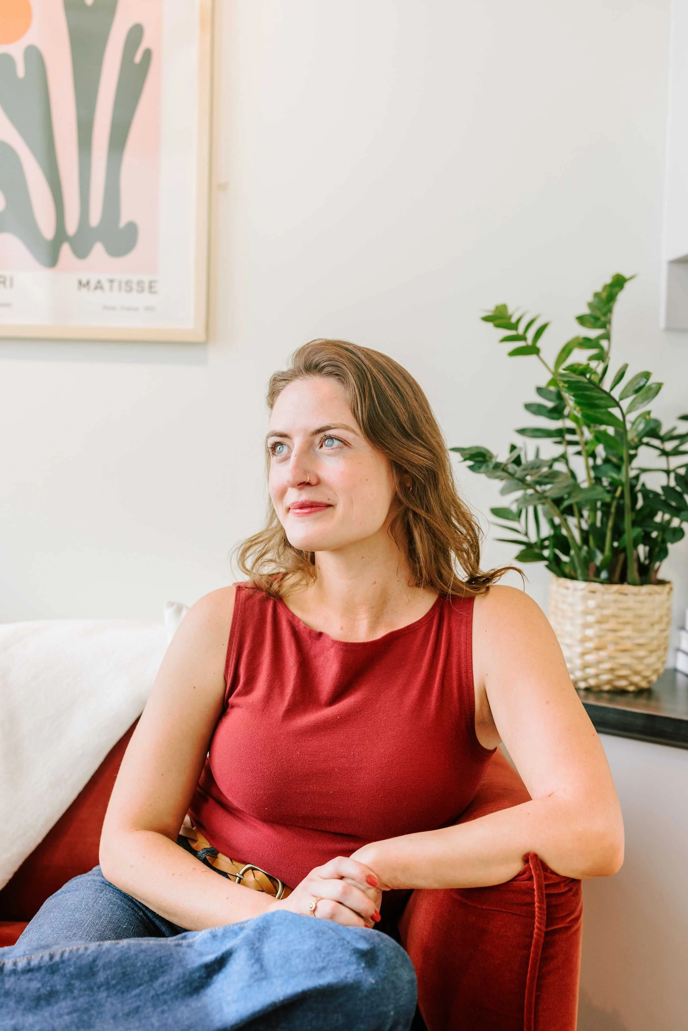 A woman with wavy brown hair and blue eyes sitting on a red couch, wearing a sleeveless red top and dark jeans, in a well-lit room with a framed artwork and a green potted plant in the background.