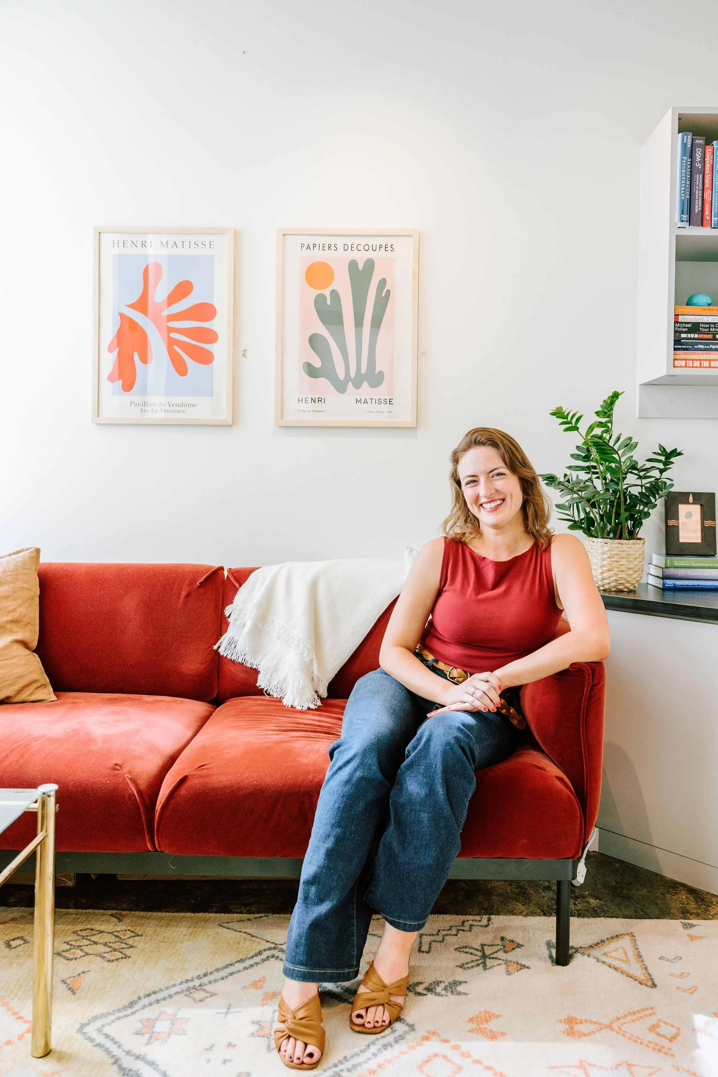 A woman sitting on a red sofa in a living room, smiling, with framed artwork on the wall behind her and a potted plant on a side table to her right.