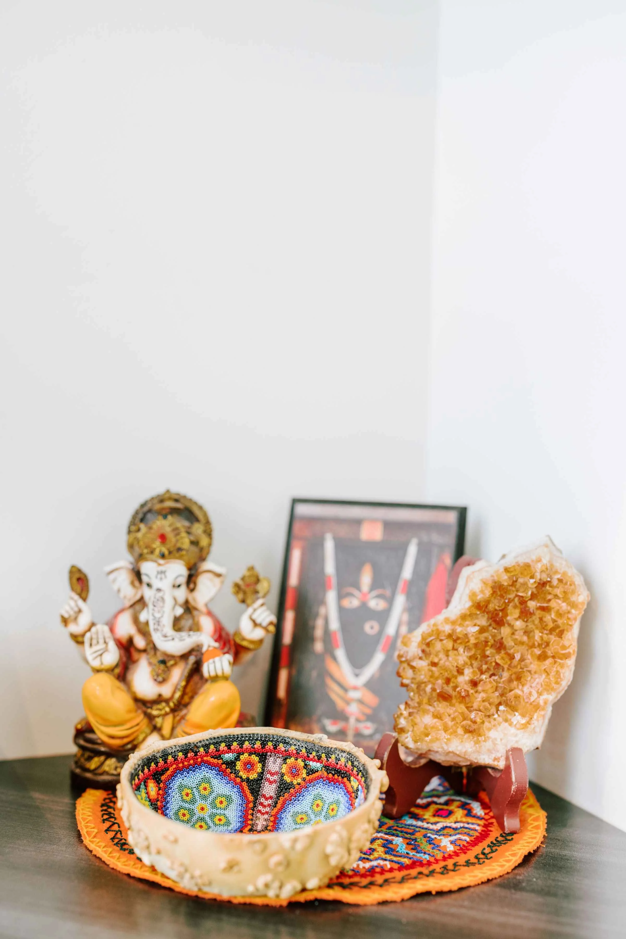 A small Hindu altar with a Ganesha idol, a beaded wristband, a framed picture of a deity, and a large orange salt crystal on a stand, all placed on a colorful mat.
