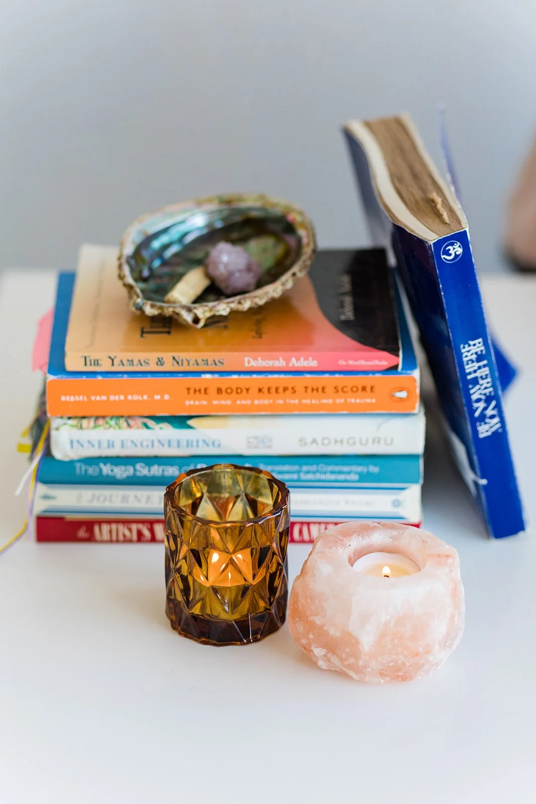 Stack of books with candles and salt lamp on a white surface, with shell and crystals on top of the books.