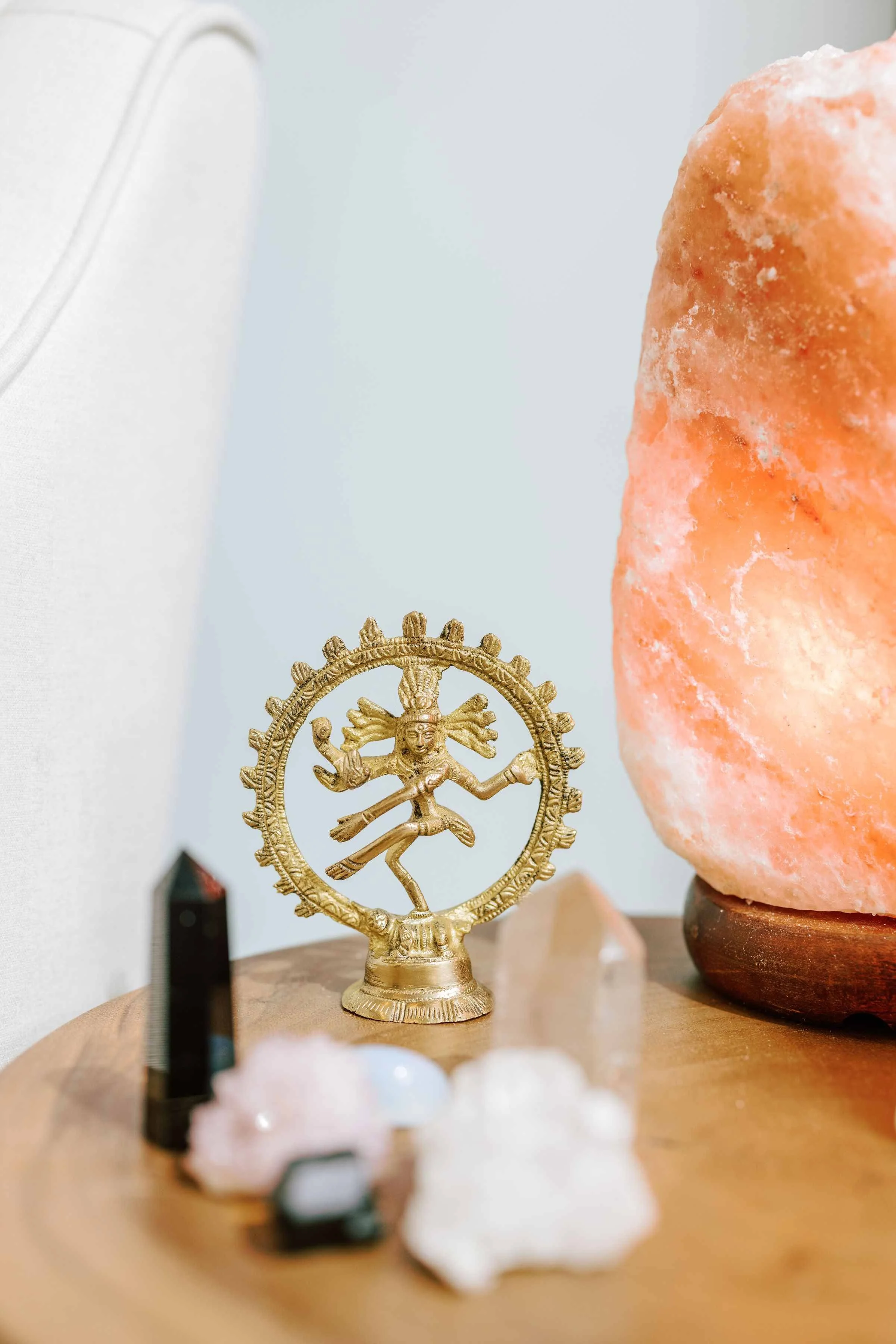 A close-up of a small golden Hindu deity statue, surrounded by various crystals and rocks on a wooden surface, with a large pink Himalayan salt lamp in the background.