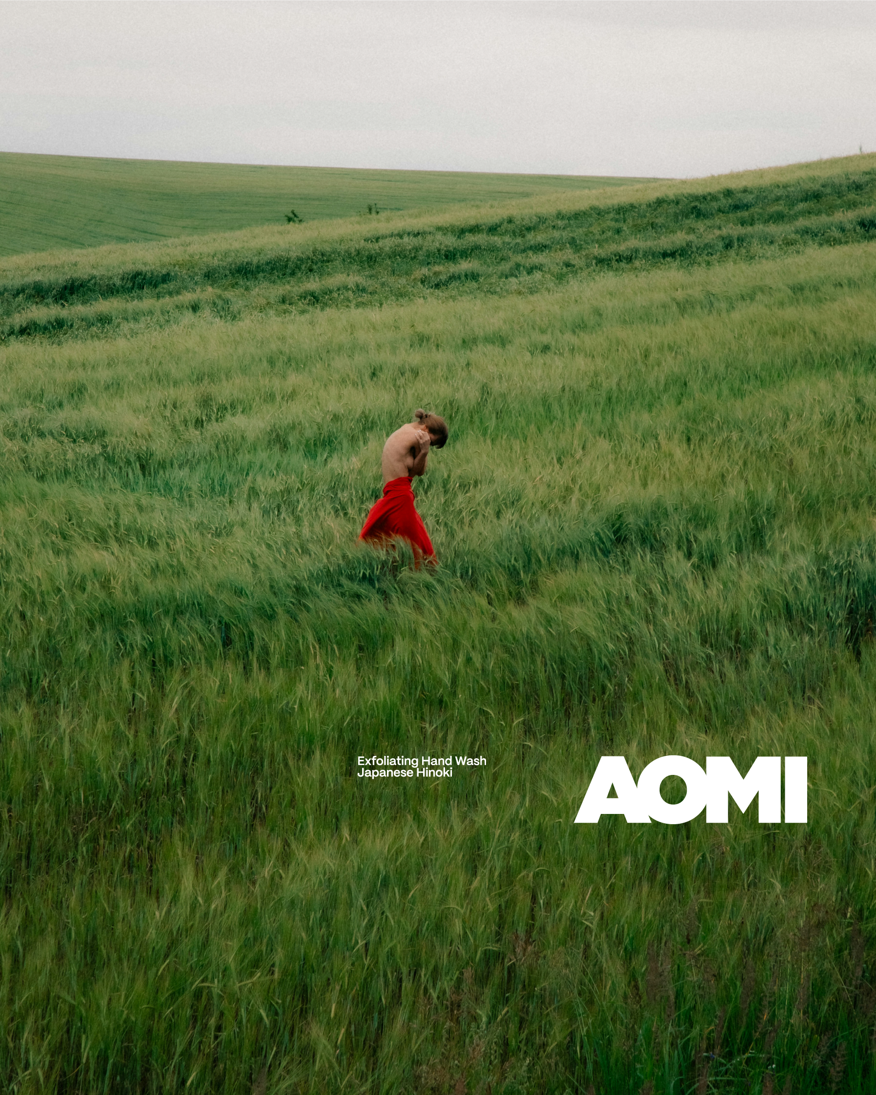 Ein Mädchen in einem roten Kleid läuft barfuß durch ein grünes Feld, im Hintergrund ein Himmel mit Wolken. Es gibt weißen Text auf dem unteren Teil des Bildes, der 'AOMI' sagt, zusammen mit klein gedrucktem Text 'Exfoliating Hand Wash Japanese Hinoki'.