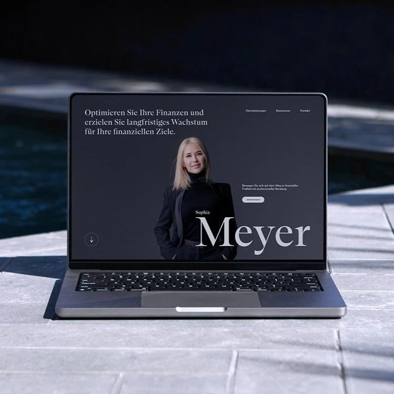 Ein Laptop mit einer Website, die eine Frau in einem schwarzen Anzug zeigt, im Hintergrund Wasser und ein Himmel, vermutlich am See oder Meer.