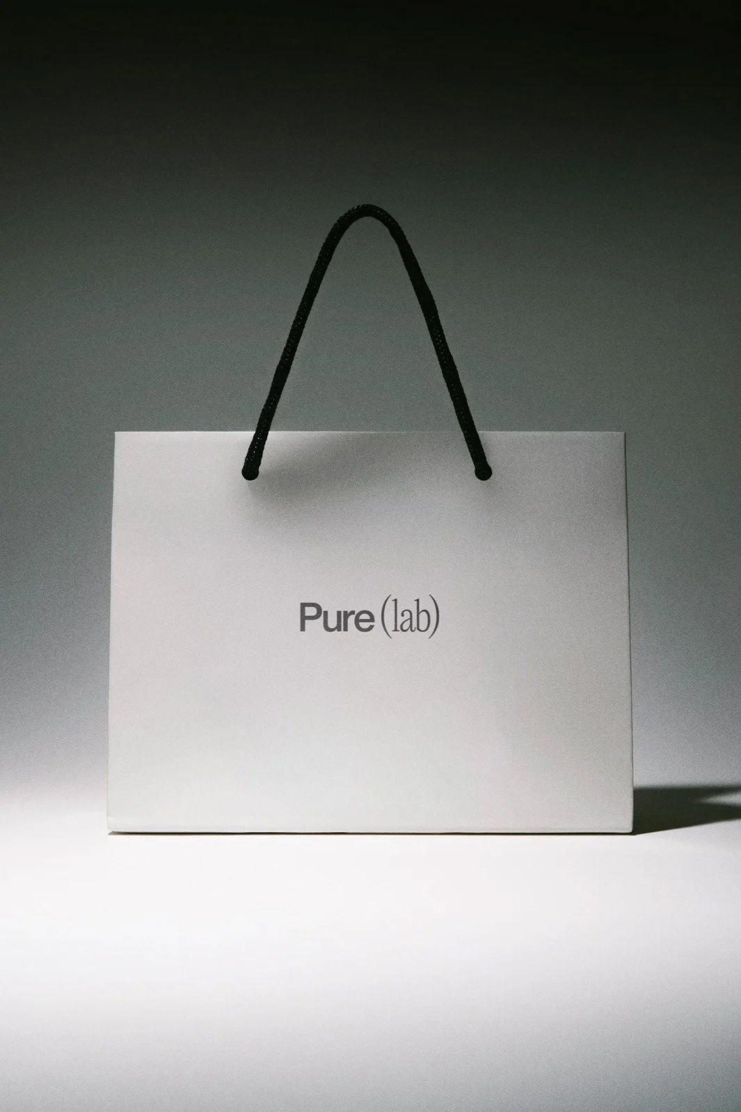 Hellgraue Einkaufstasche mit der Aufschrift "Pure(lab)" hängt an einem Türgriff neben einer Holzwand und einem weißen Türrahmen.