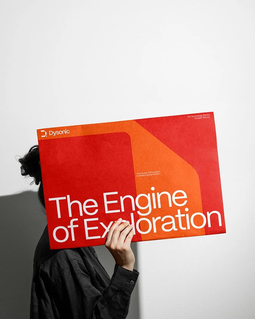 Person hält ein rotes Buch mit dem Titel 'The Engine of Exploration' vor Gesicht, Person trägt schwarze Jacke, Hintergrund ist weiß.