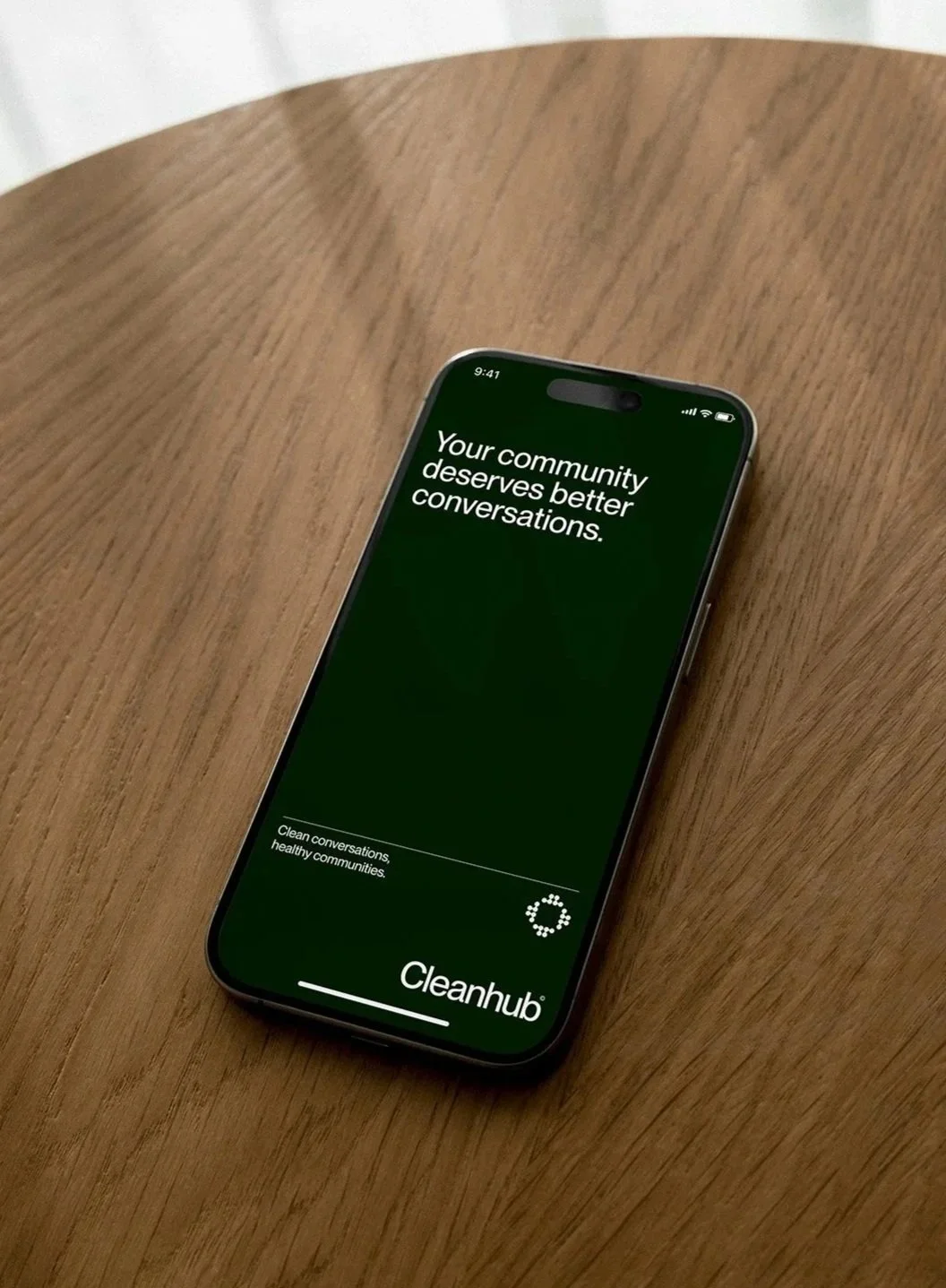 Ein Smartphone liegt auf einem runden Holztisch. Auf dem Bildschirm ist die App Cleanhub geöffnet, die den Slogan 'Your community deserves better conversations' zeigt.