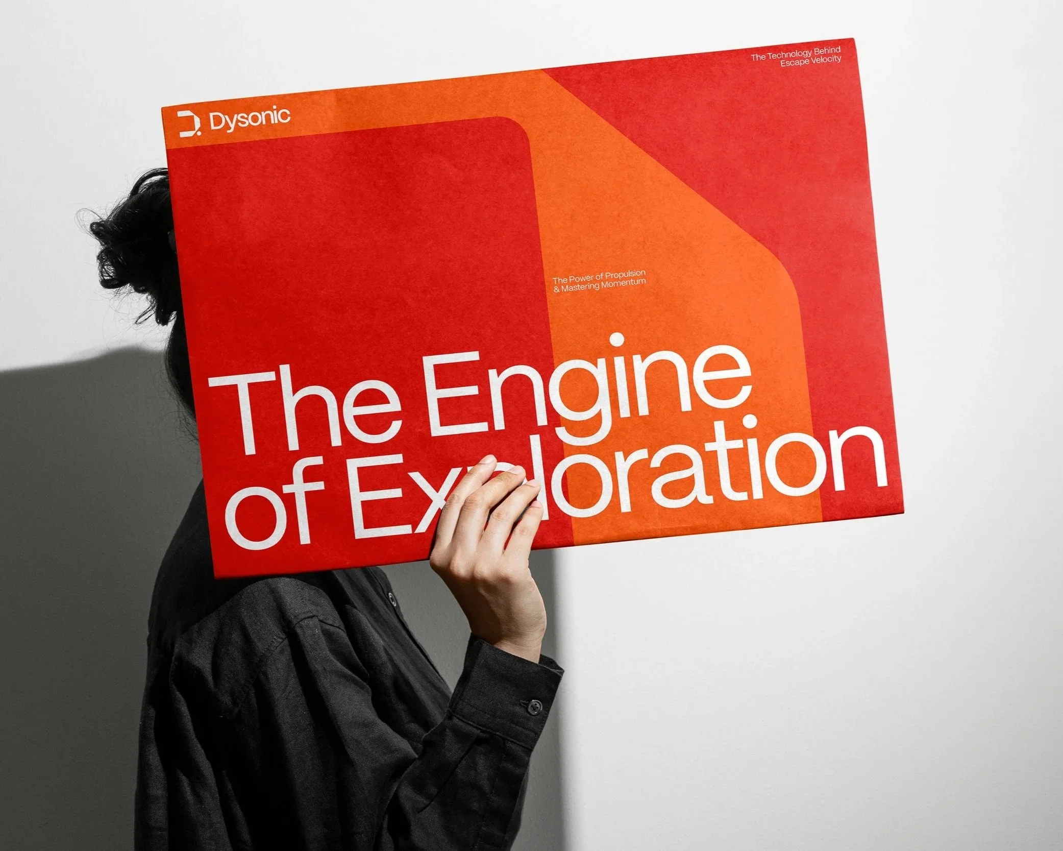 Person hält ein rotes Buch mit dem Titel 'The Engine of Exploration' vor Gesicht, Person trägt schwarze Jacke, Hintergrund ist weiß.