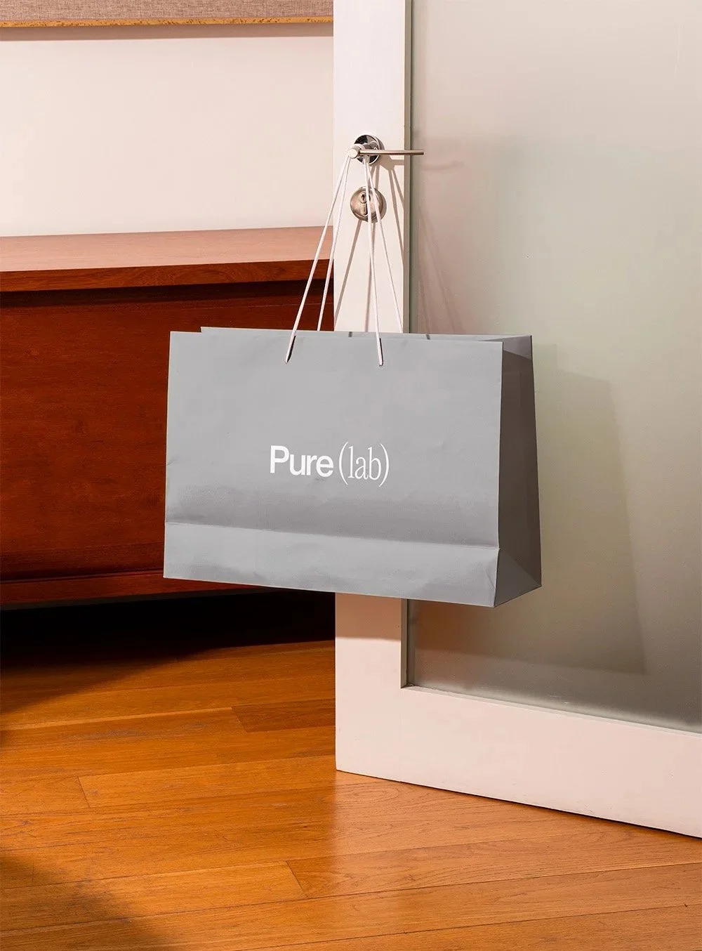 Hellgraue Einkaufstasche mit der Aufschrift "Pure(lab)" hängt an einem Türgriff neben einer Holzwand und einem weißen Türrahmen.