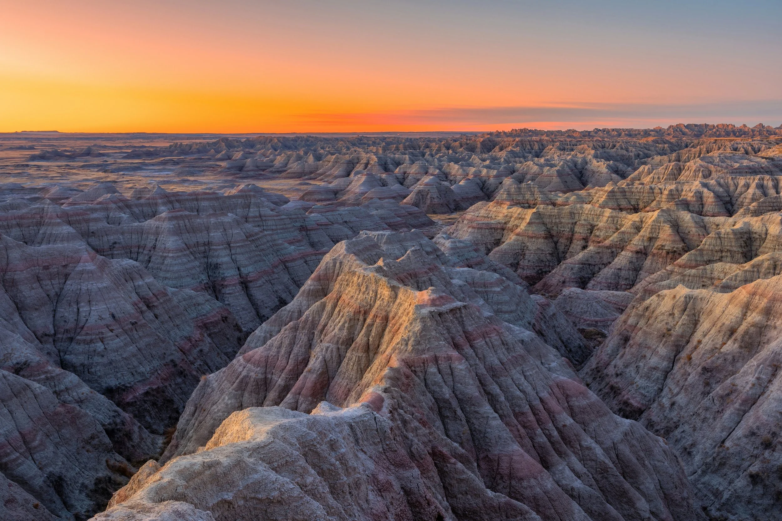 The image shows a vast landscape of rugged, eroded rock formations at sunset, with a colorful sky featuring shades of orange, pink, and blue.