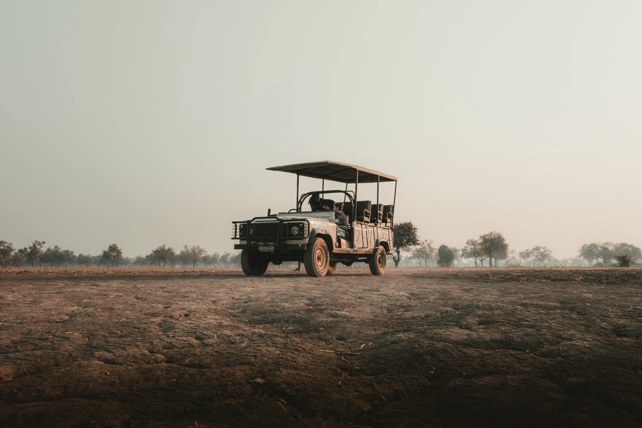 An open safari vehicle parked on a dirt field with sparse trees in the distance under a cloudy sky.