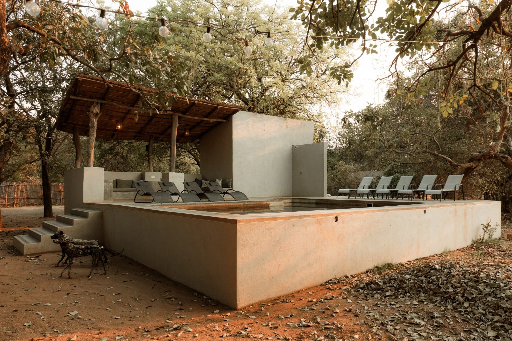 An outdoor pool area with surrounding lounge chairs, trees with hanging string lights, and a shaded seating area with cushions.