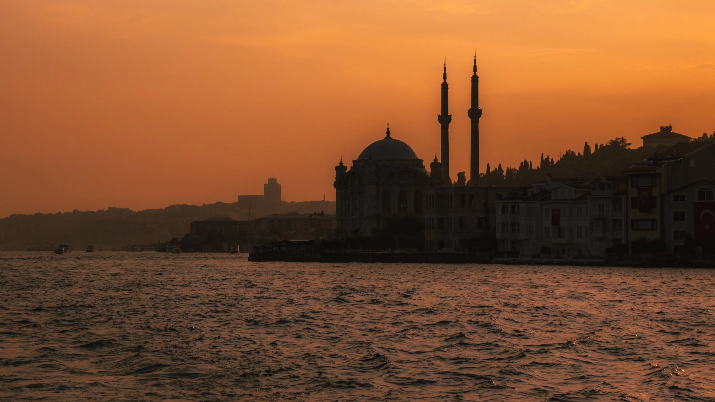 Sunset over a body of water with boats, with a silhouette of a mosque with twin minarets and buildings along the shoreline.