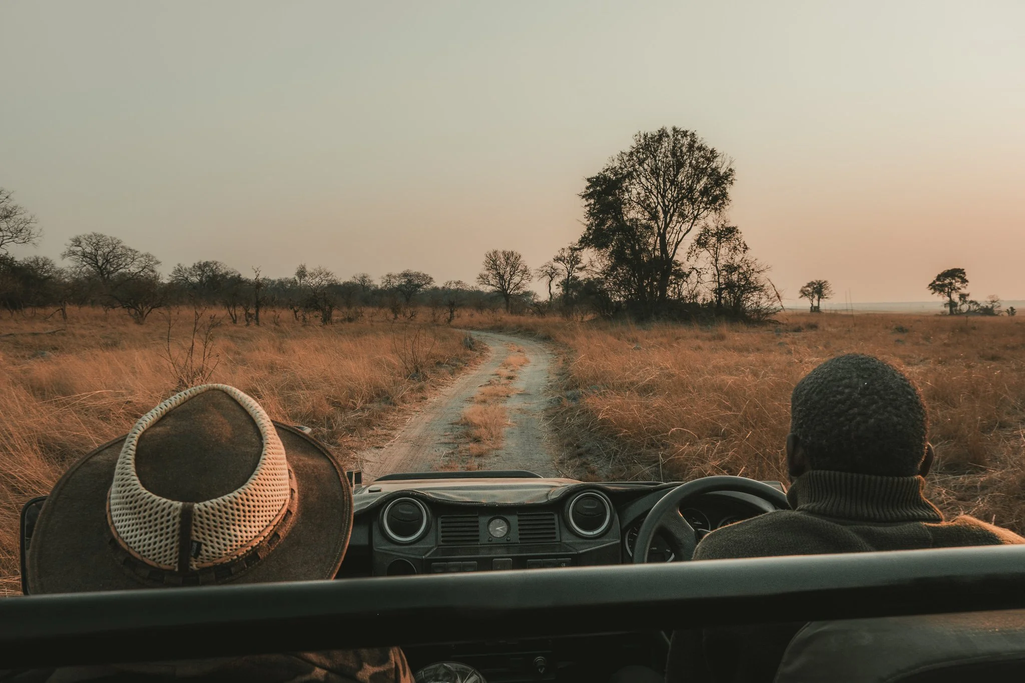 View from inside a vehicle driving on a dirt road through a grassy savannah landscape with sparse trees at sunset, with two people in the front seats, one wearing a hat and the other with short curly hair.