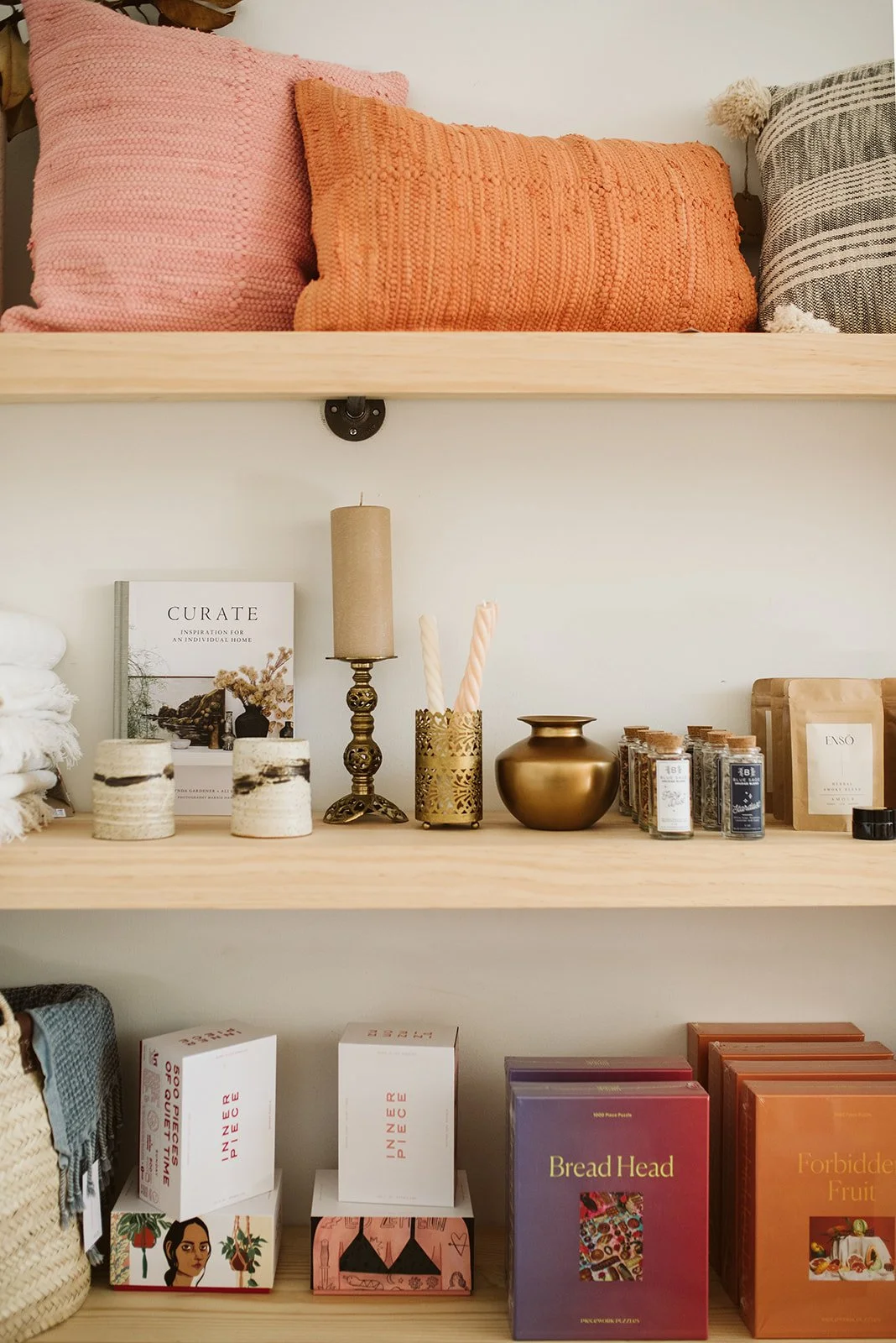 Decorative shelf displaying pillows, a book titled 'Curate,' a beige candle, a ornate metal candle holder with two pink swirled candles, a metallic gold vase, some small jars, and various books and packaging.