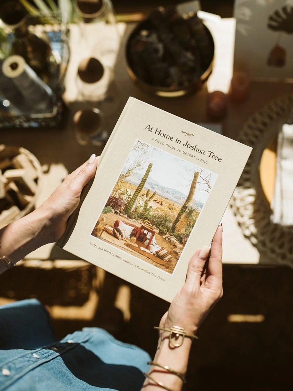 A person holding a book titled 'At Home in Joshua Tree: A Field Guide to Desert Living' with an image of a desert scene featuring cacti on the cover. The background shows a cluttered workspace with various objects.