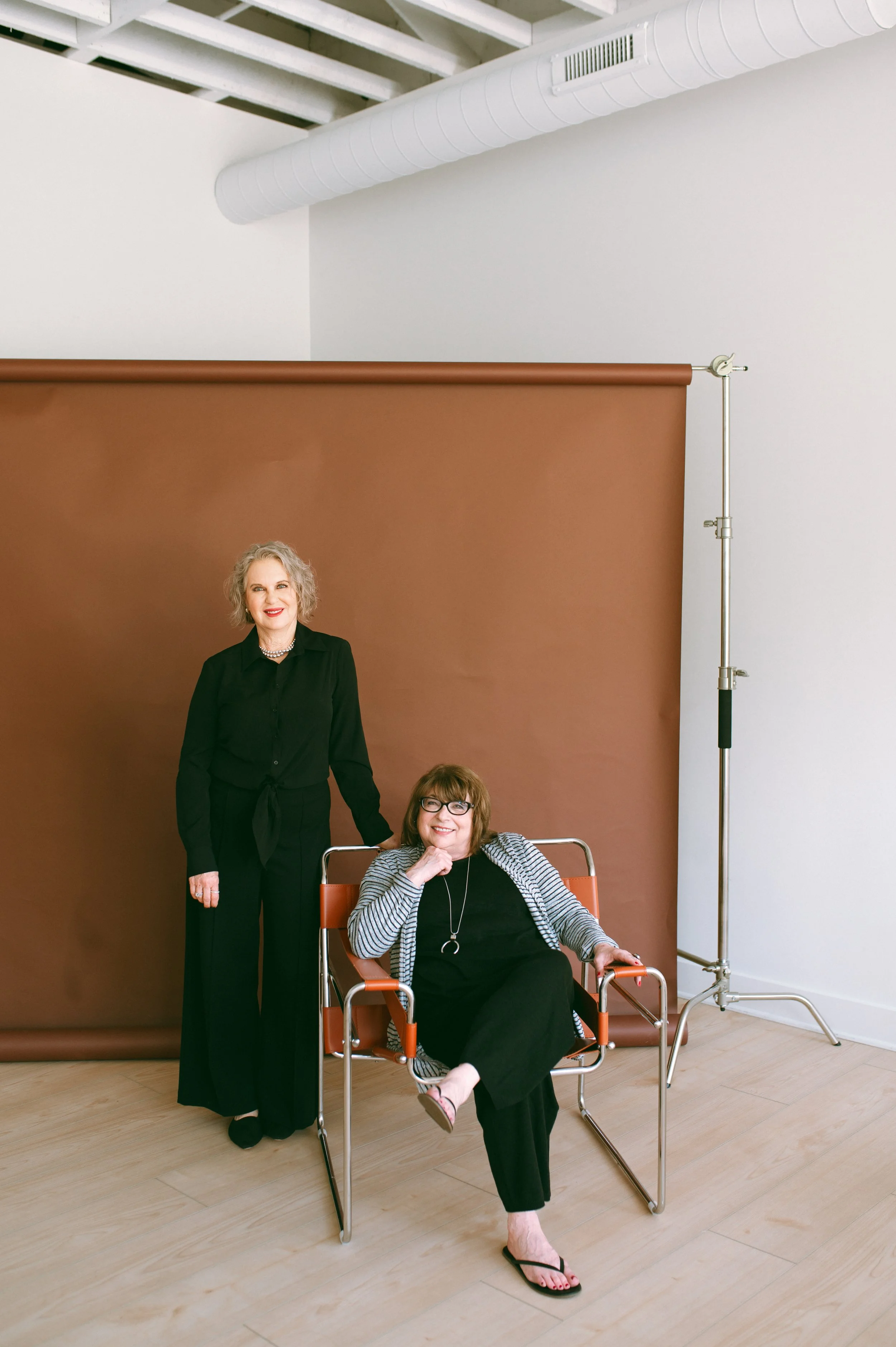 Two women in a photography studio with a brown backdrop. One woman, with curly gray hair and wearing black, is standing and holding the hand of a seated woman with glasses and wearing a striped jacket. The seated woman is smiling, with one leg crosse