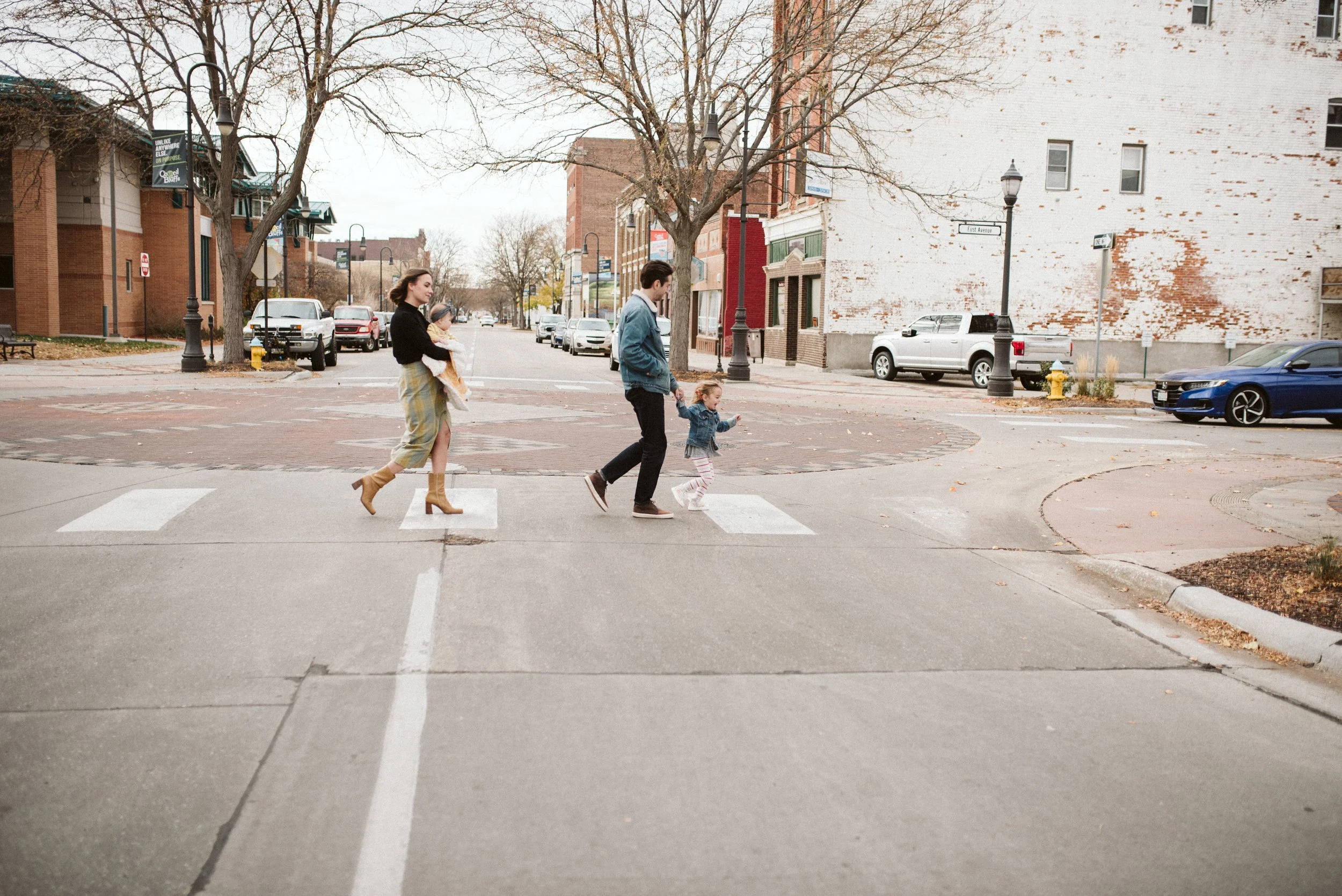 A family crosses a city street at a crosswalk, with a woman holding a child, a man holding a young girl, and cars parked along the street, during autumn with bare trees and falling leaves.
