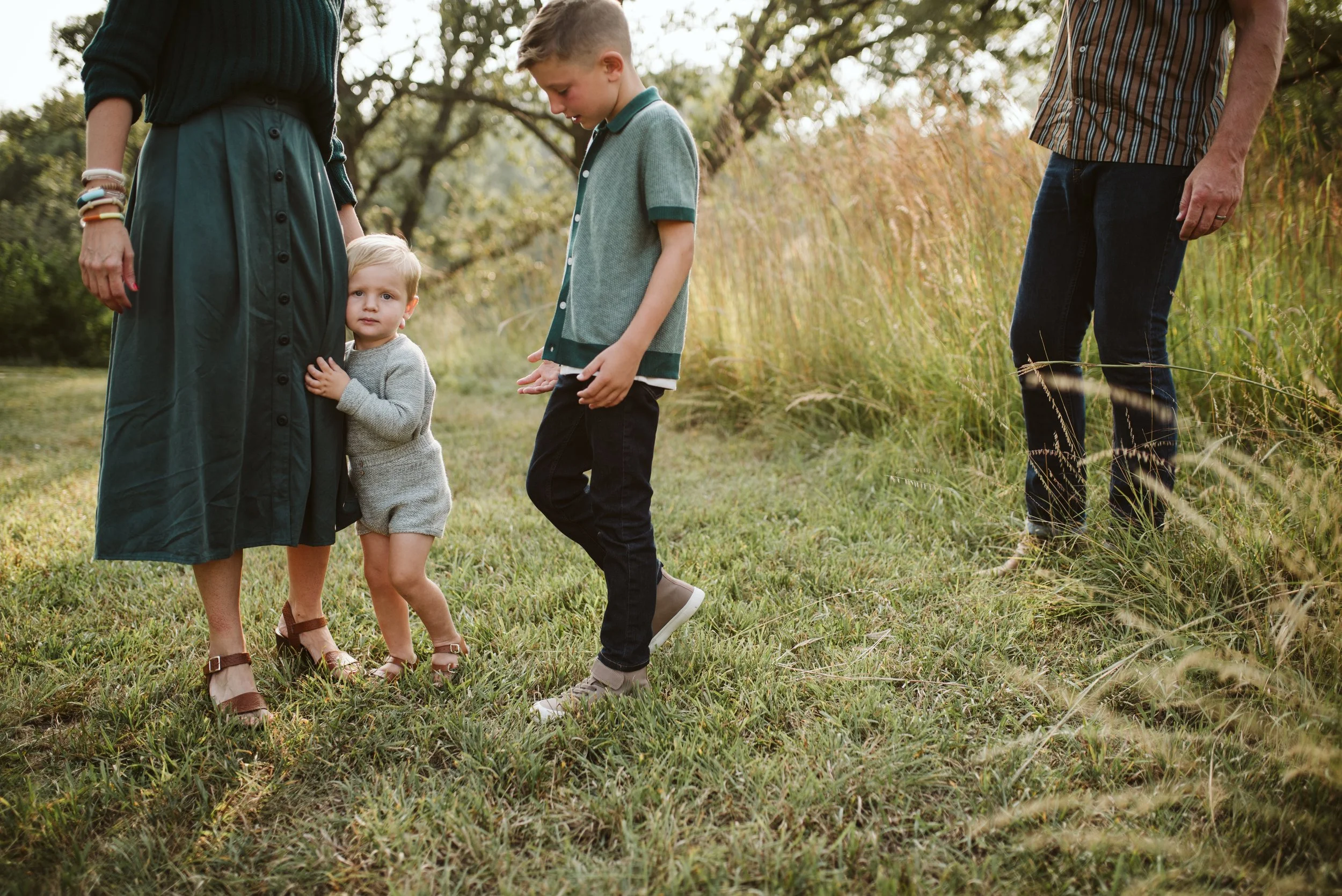 A family with two children standing outdoors in a grassy area surrounded by trees, with the children looking toward the camera.