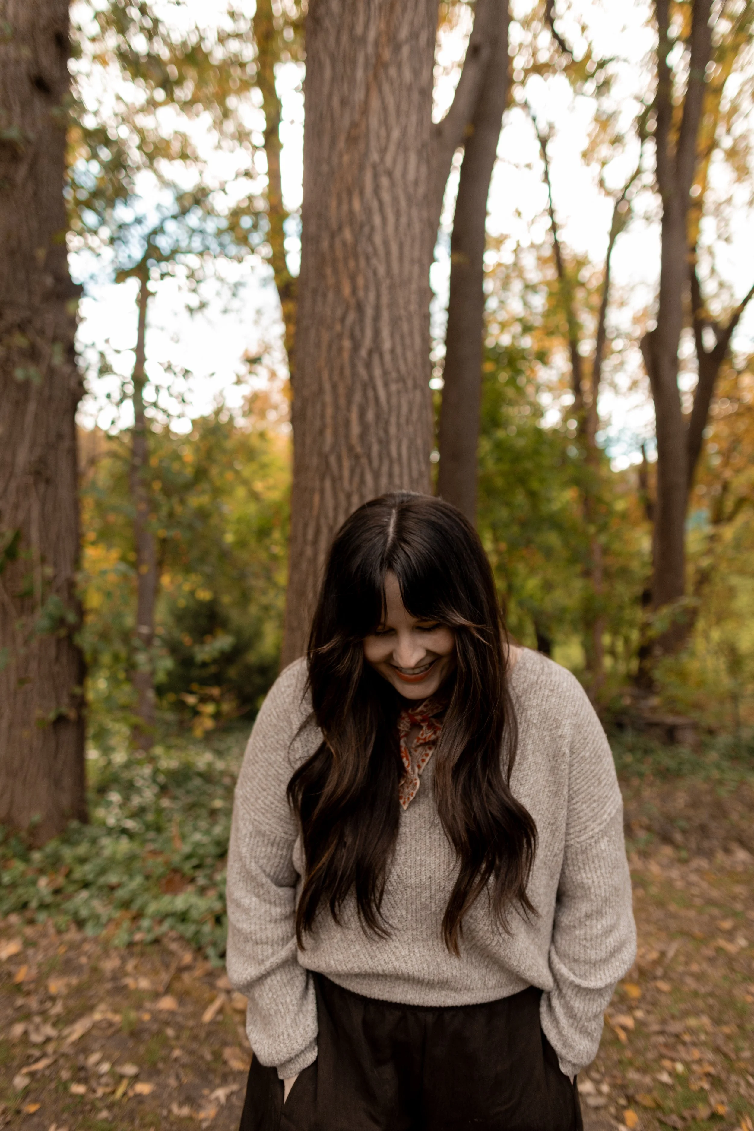 A woman with long dark hair smiling and looking down in a forest with tall trees and autumn leaves.