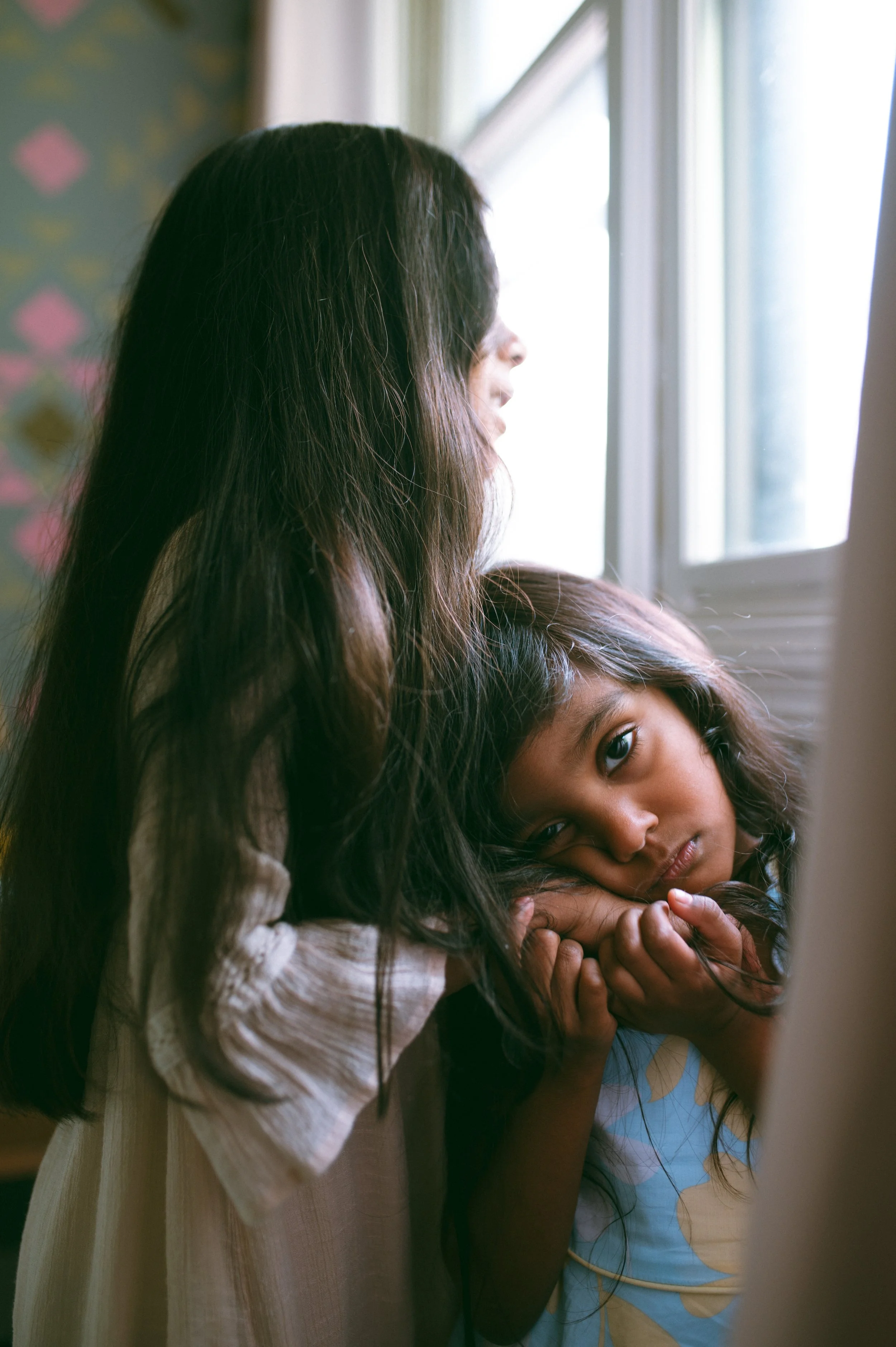 A woman with long dark hair looking out a window, with a young girl resting her head on her arm, looking at the camera, in a softly lit room.