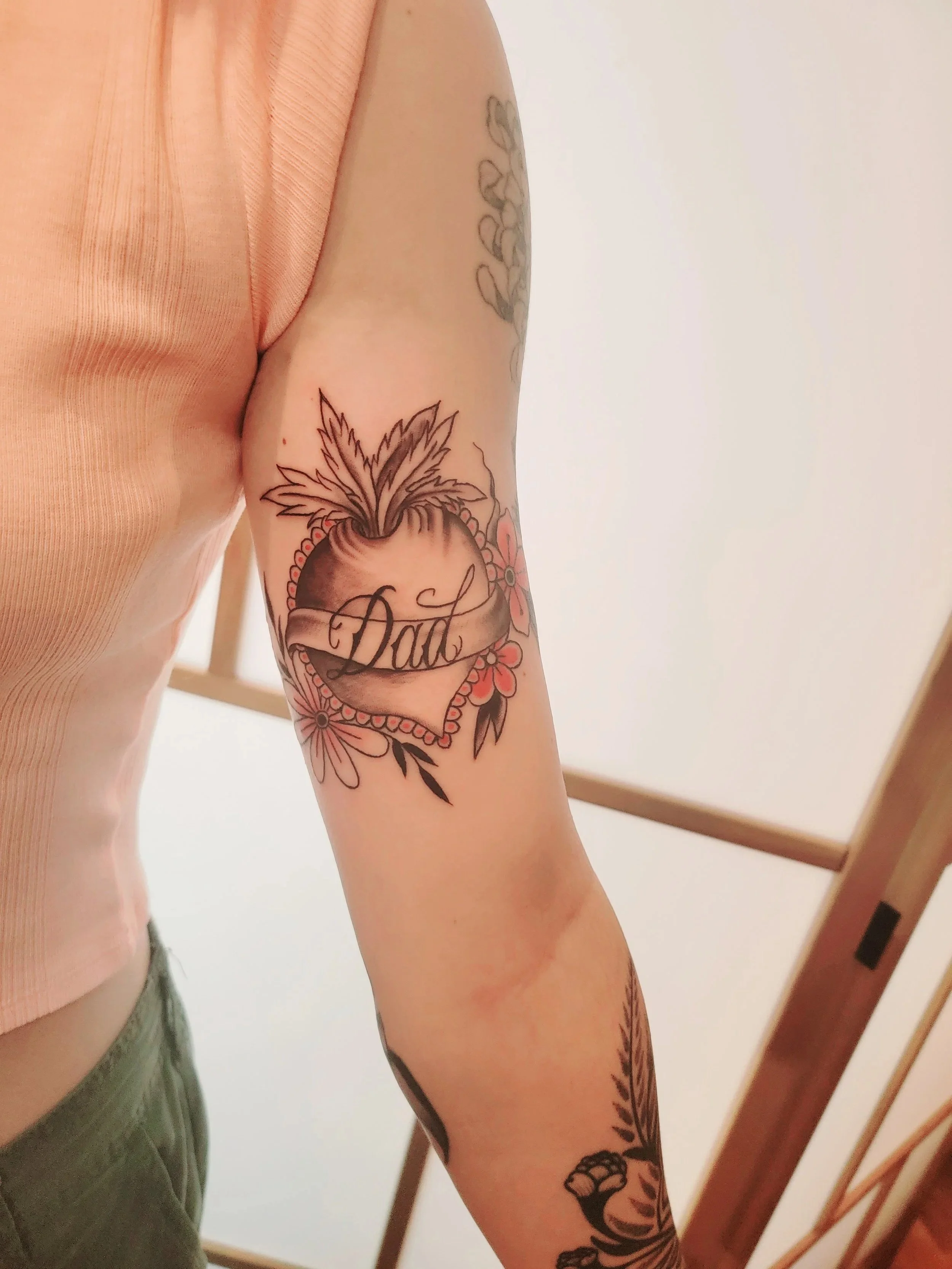 Tattoo of a heart with banner reading "Dad", surrounded by flowers, on person's arm.