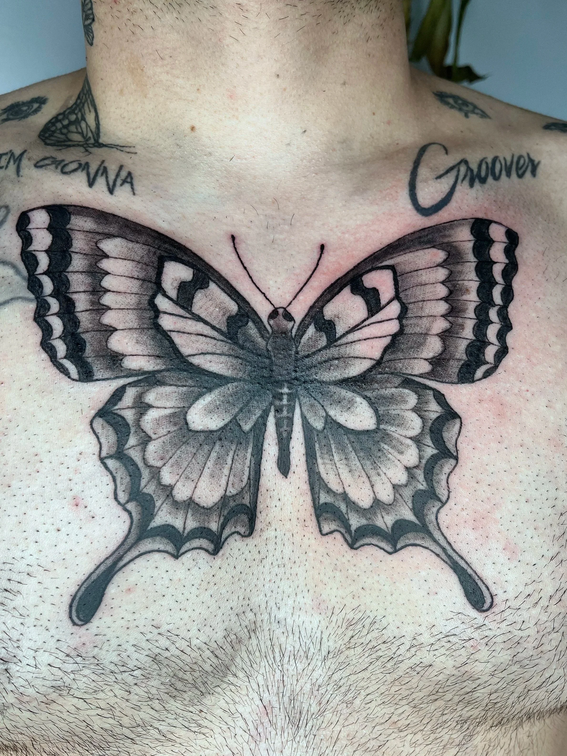 A tattoo of a butterfly on a person's chest, with tattooed words 'Groover' and 'I'm Gonna' on the collarbone area.