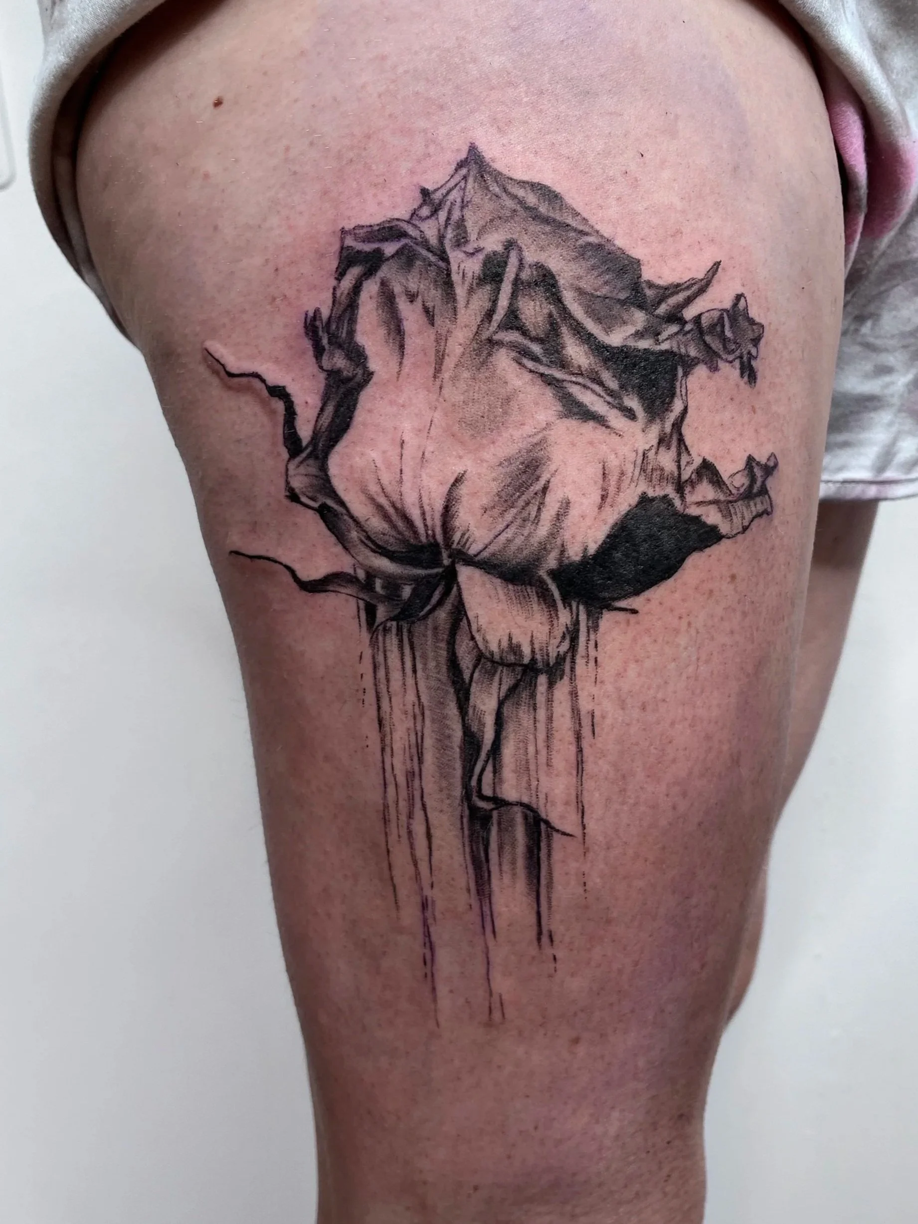 Black ink tattoo of a rose on a person's upper thigh with sketchy, watercolor-style shading and dripping effects.