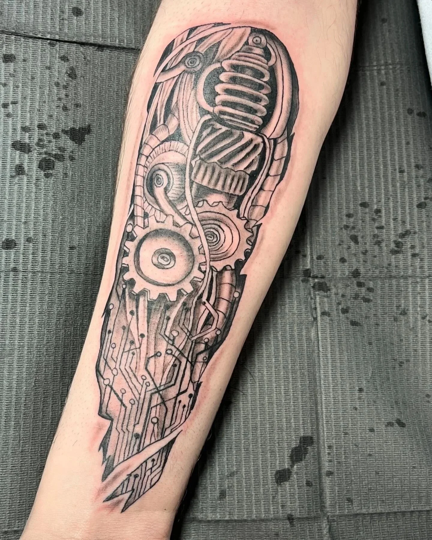 Black and grey tattoo of a mechanical and circuit theme, featuring gears, cogs, and circuit lines, on a person's forearm.