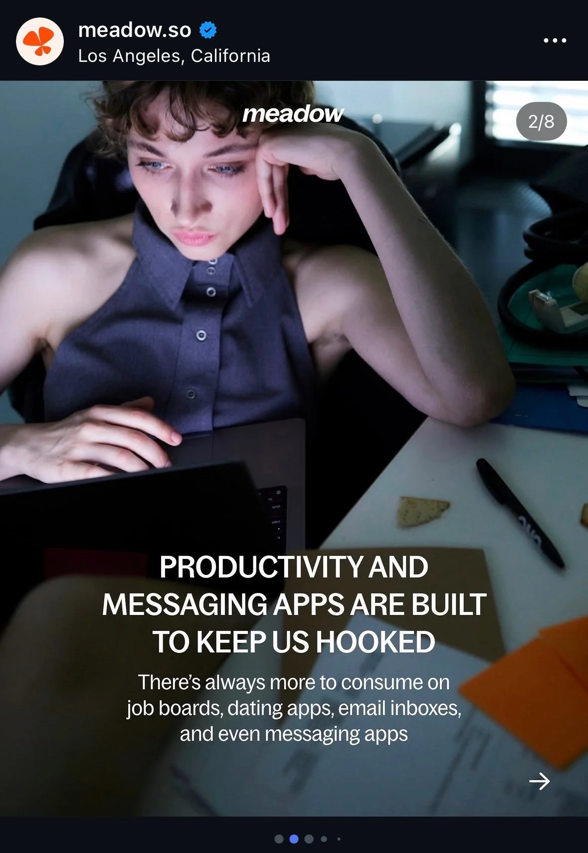 Young person with short curly hair sitting at a desk, looking at a laptop, with a focused expression.