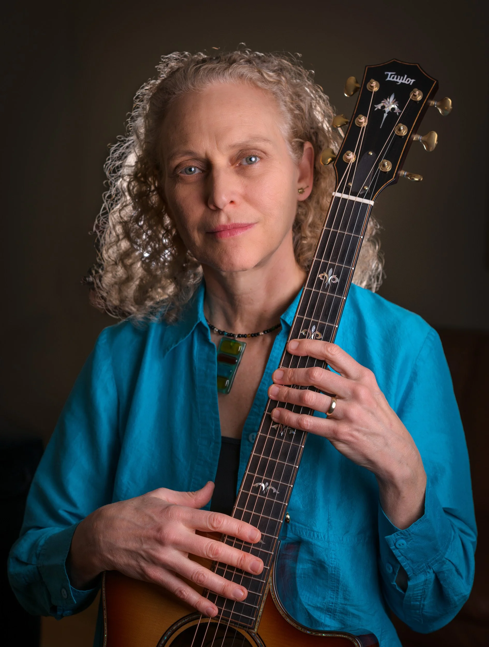 Portrait photo of Guitarist Mimi Fox wear a blue shirt and holding a six-string acoustic guitar.
