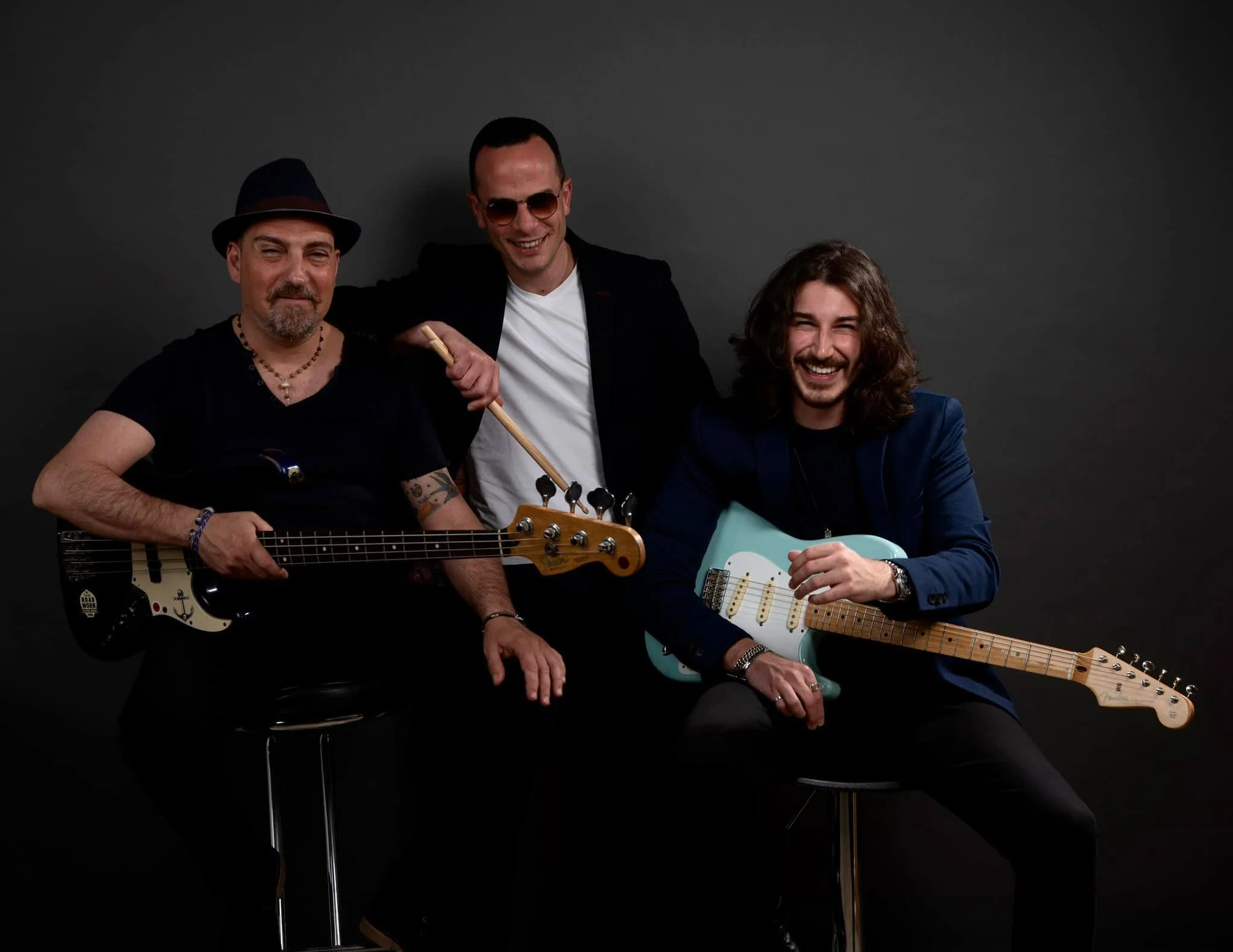 Photo of three band members posing with electric bass guitar, drum sticks, and electric guitar.