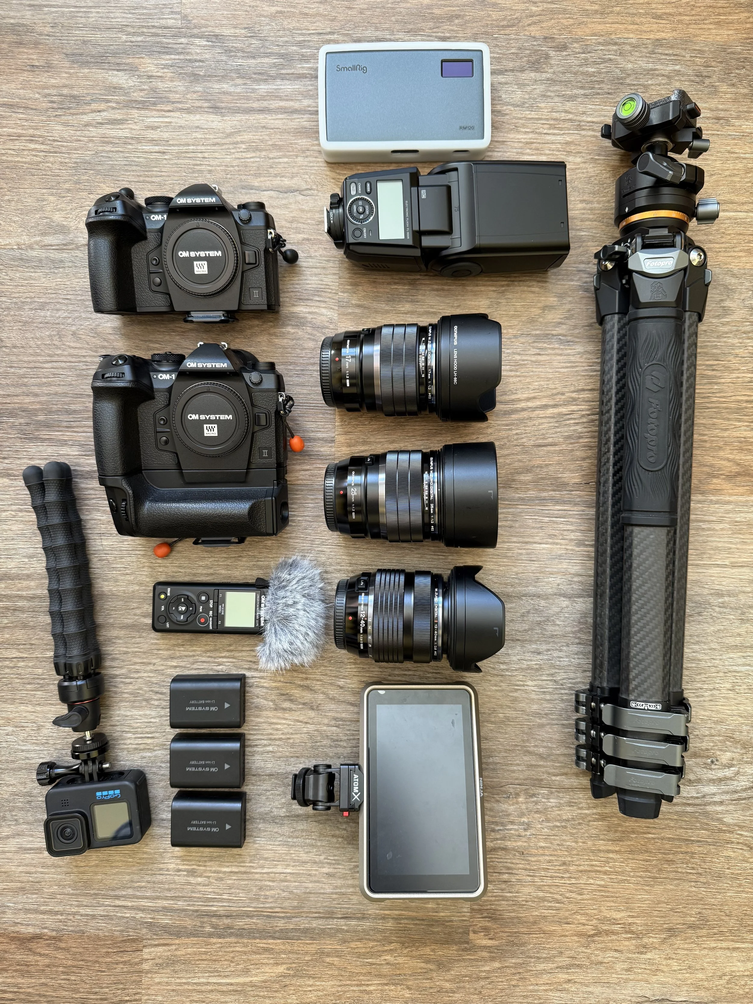 Two OM-1 Mark ii cameras, OM 25mm f/1.2, OM 17mm f/1.2, OM 12-40mm f/2.8, assorted batteries and camera accessories laid out on ground ready to be packed.
