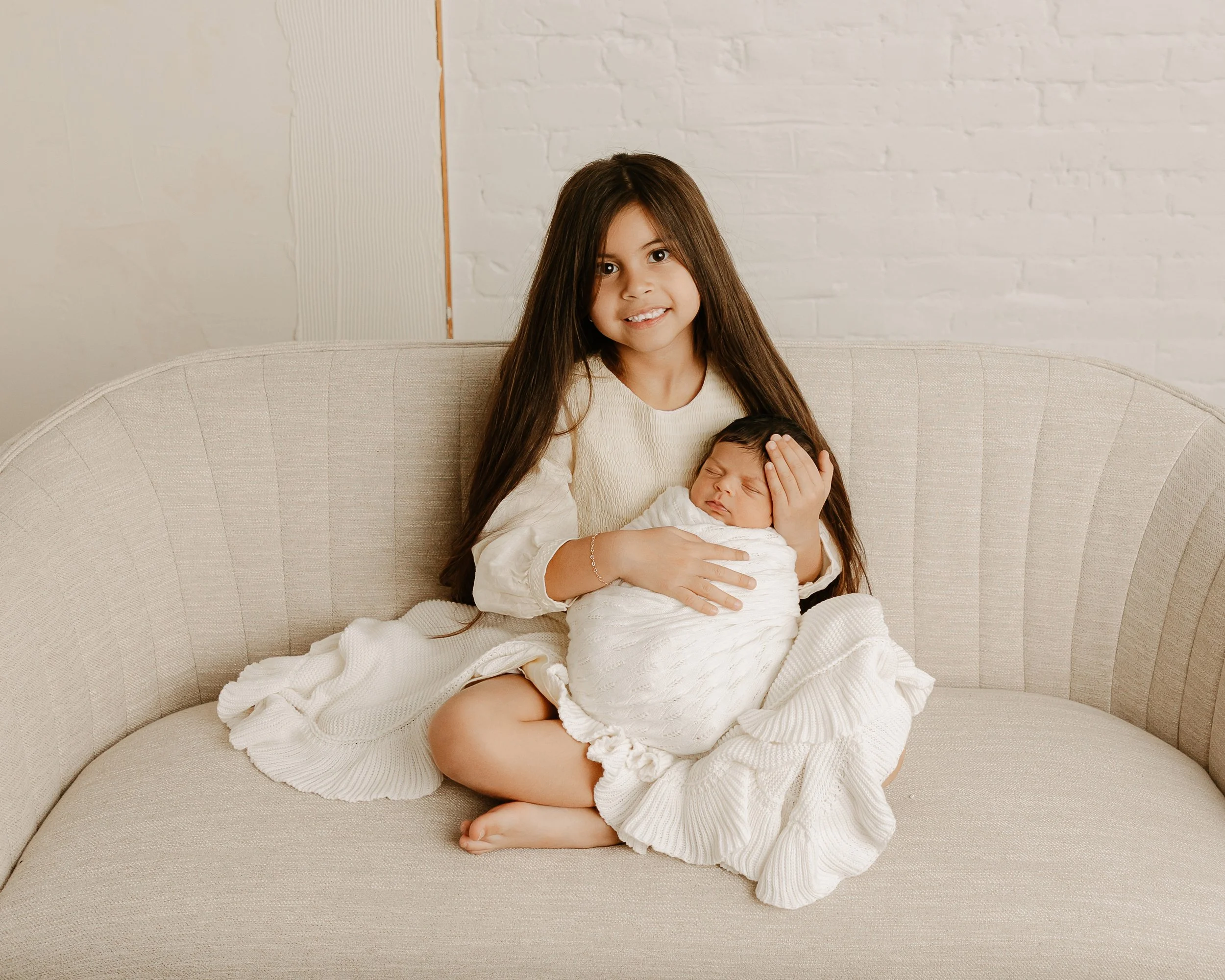 older sister holds her newborn baby sister wrapped in a white blanket on a comfy couch during a newborn photoshoot
