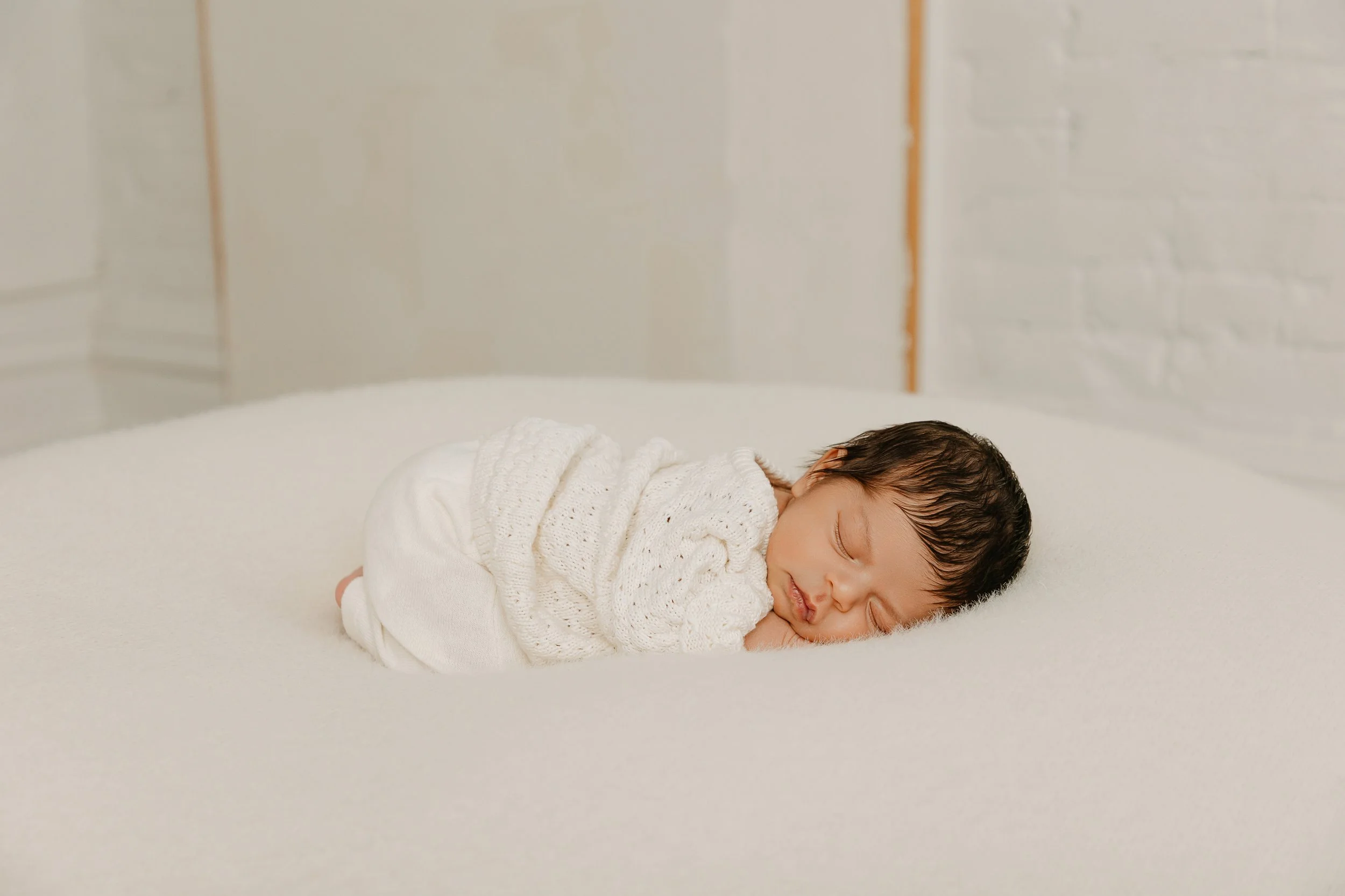 newborn baby in a textured knit outfit sleeps on a soft white pillow in a white room