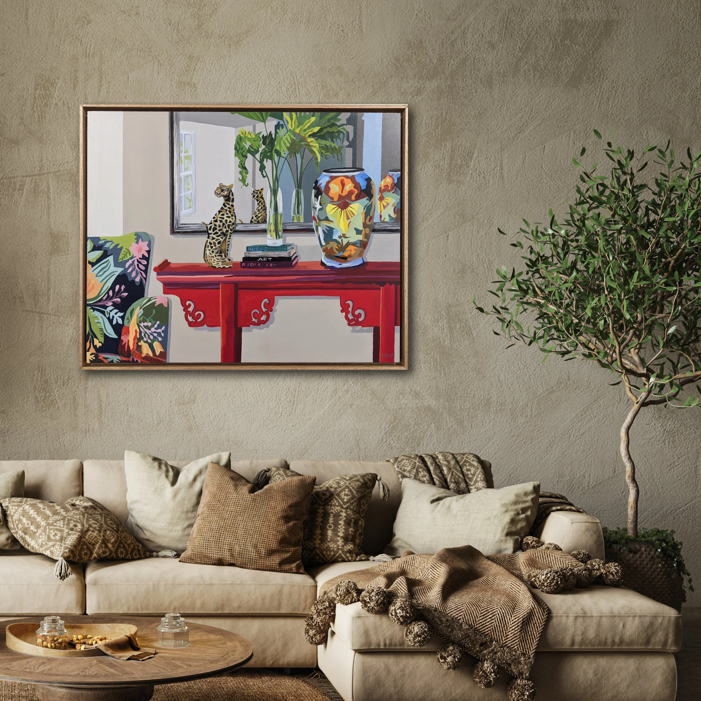 Living room with beige sofa, decorative pillows, a textured knit throw, and a round wooden coffee table with snacks. A framed painting above the sofa depicts a room with a colorful chair, red side table, books, a plant, and a decorative vase. A potted green plant is beside the sofa.