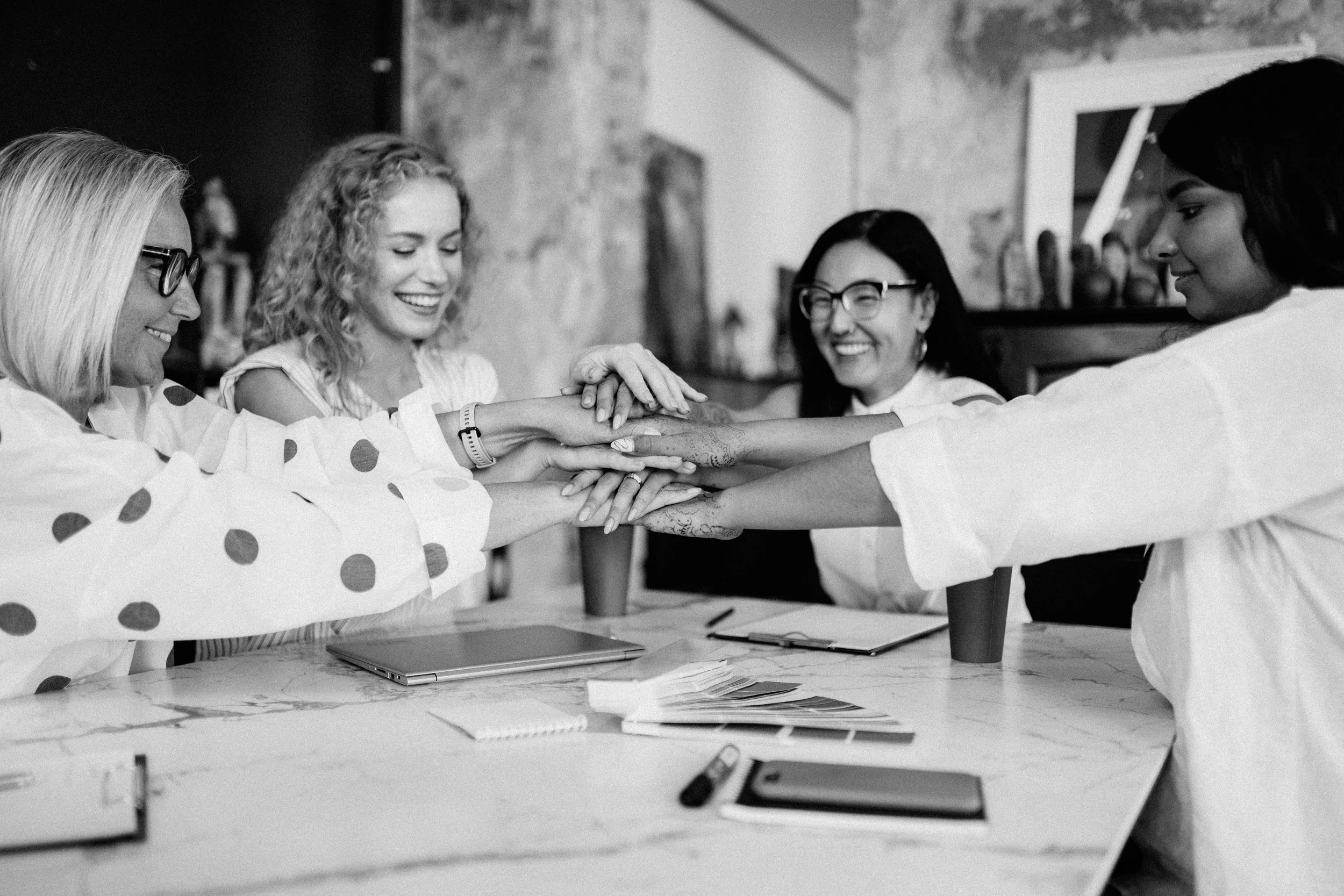 Five women in an office sitting around a table, stacking their hands together in a show of teamwork and friendship, smiling and appearing joyful.