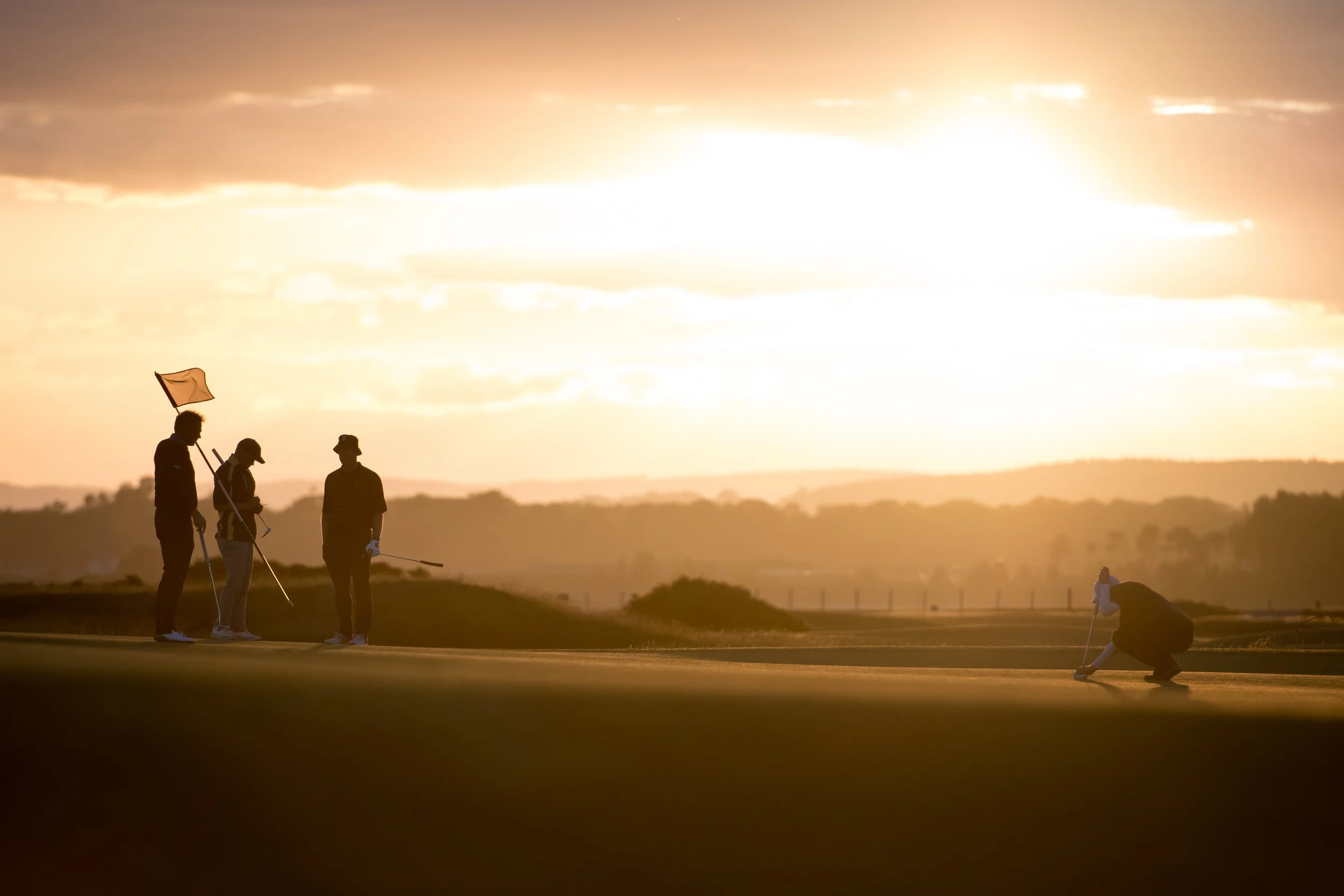 Four people playing golf on a course during sunset, with one putting and the others watching, silhouetted against a bright sky.