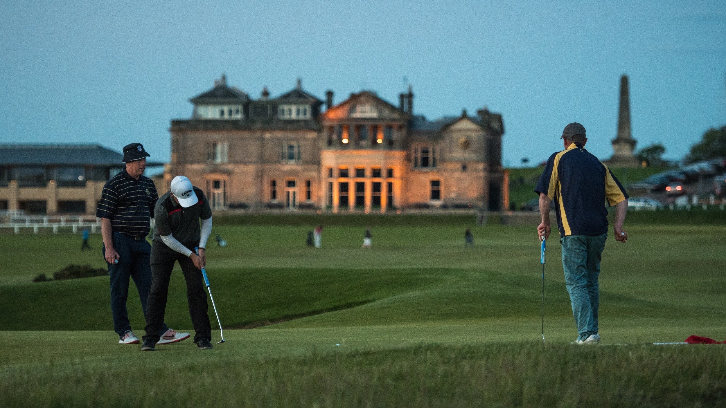 Three men playing golf on a green, with a large historic building in the background during twilight.