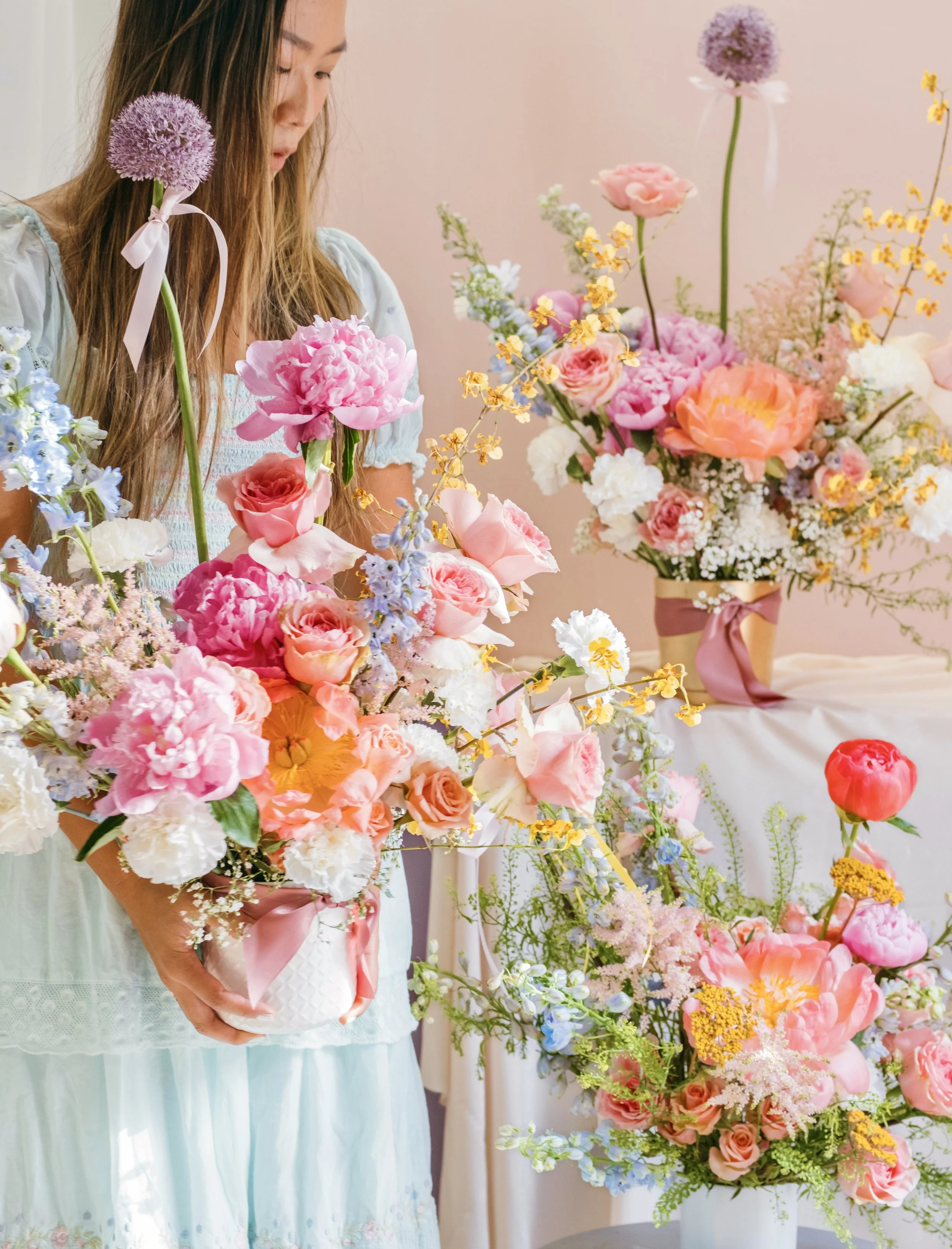 A woman holding a bouquet of pink and white flowers in a white vase with pink ribbons, in front of more pink and white floral arrangements on a table, set against a pink background.