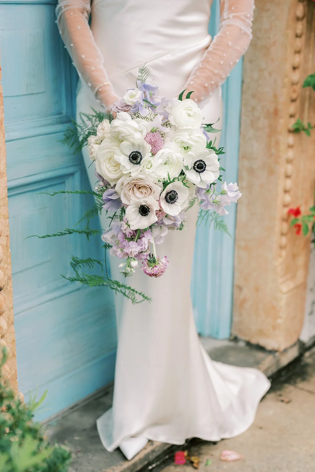 A woman in a white dress holding a cascading bouquet of white, lavender, and blush flowers with greenery, standing by a blue door.