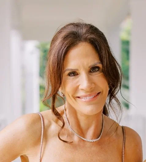 A smiling woman with brown hair and a pearl necklace, wearing a dress with spaghetti straps, standing in a bright outdoor setting.