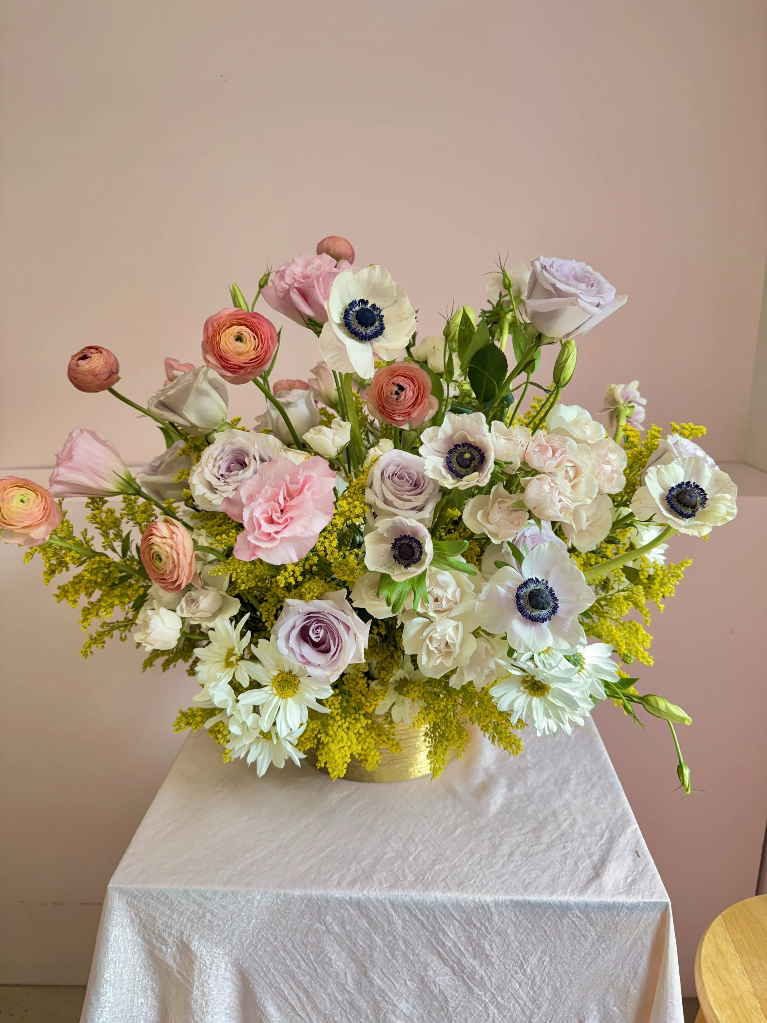 A floral arrangement with pink, purple, white, and yellow flowers in a gold vase on a white cloth-covered table.