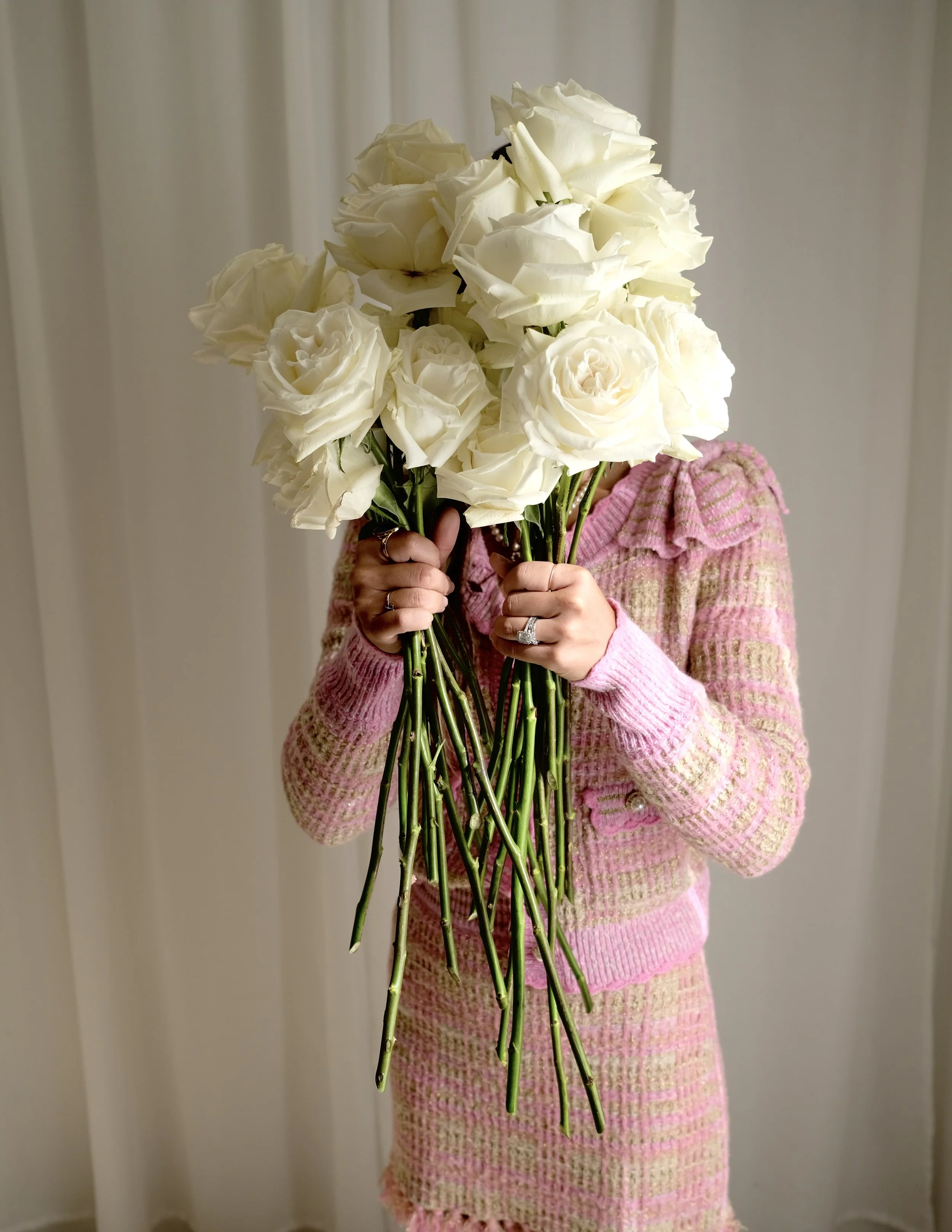 Person holding a large bouquet of white roses in front of her face, wearing a pink and beige checkered sweater and skirt, standing indoors.