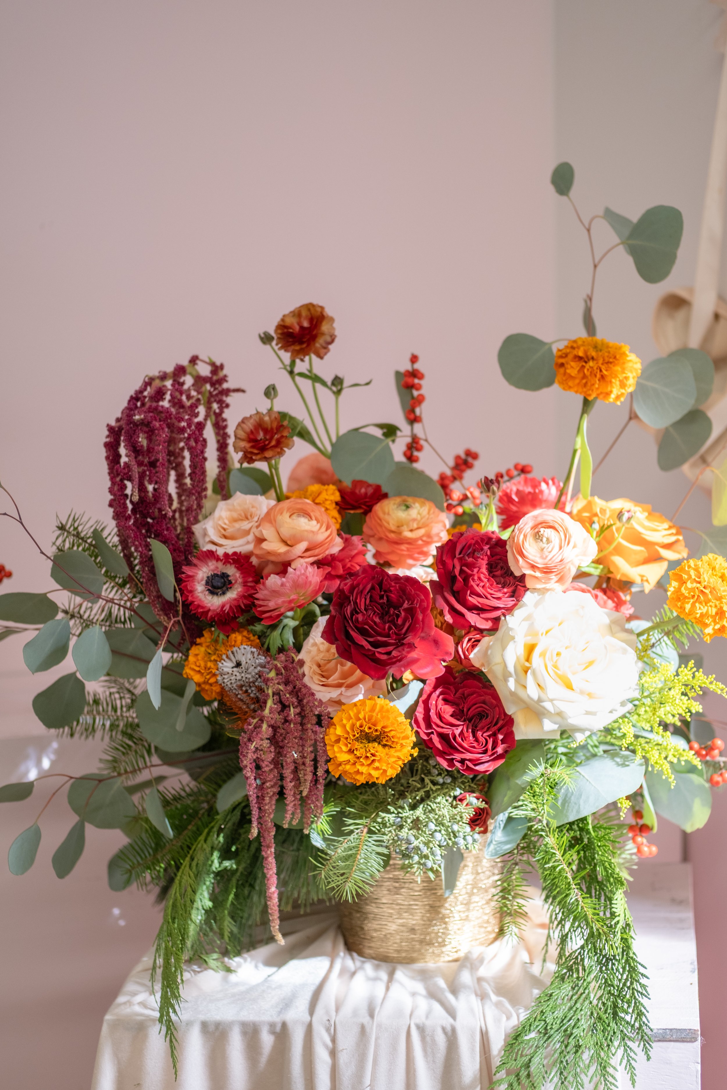 A colorful floral arrangement in a woven basket with pink, red, yellow, and white flowers, and green foliage, placed on a white cloth-covered table. Perfect Thanksgiving, Valentine's day flower.