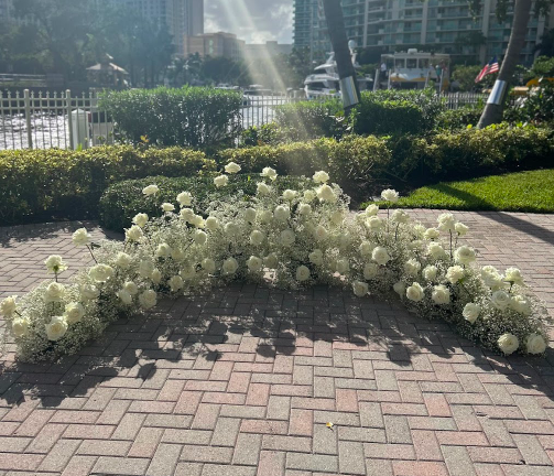 A floral arrangement of white flowers placed on a brick sidewalk outside, with a fence, shrubs, trees, and buildings in the background.