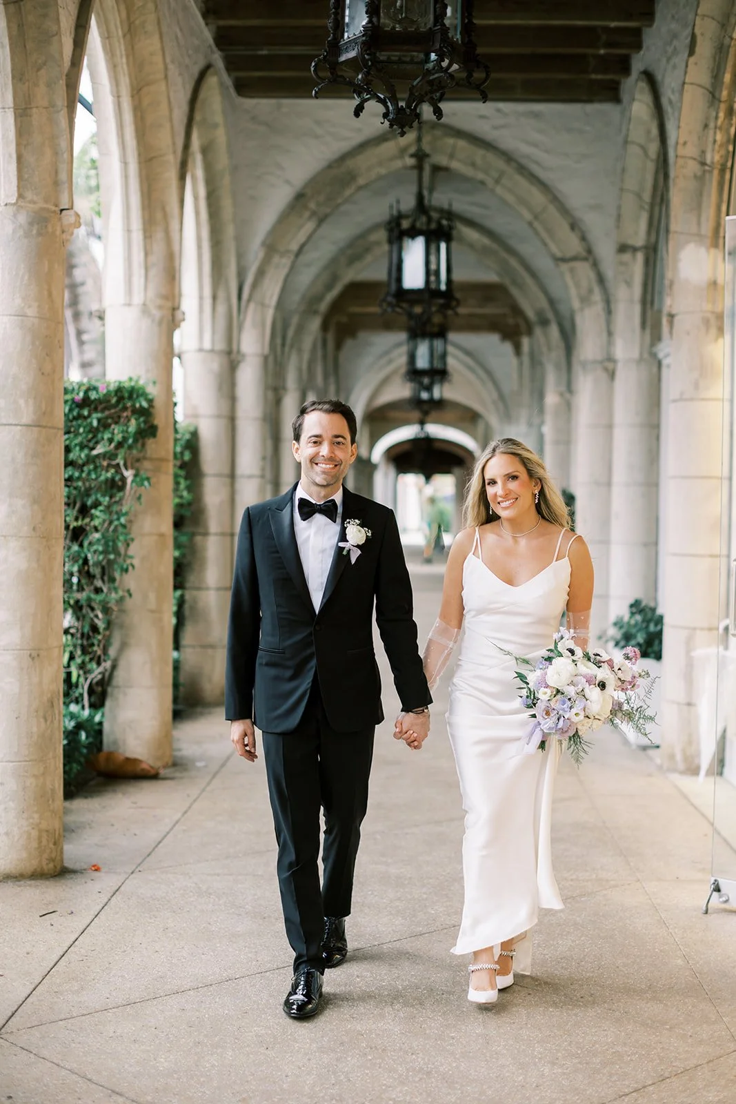 A newlywed couple walking hand in hand through an arched outdoor walkway, the groom in a black tuxedo and the bride in a white satin wedding dress holding a bouquet of white and pastel flowers.