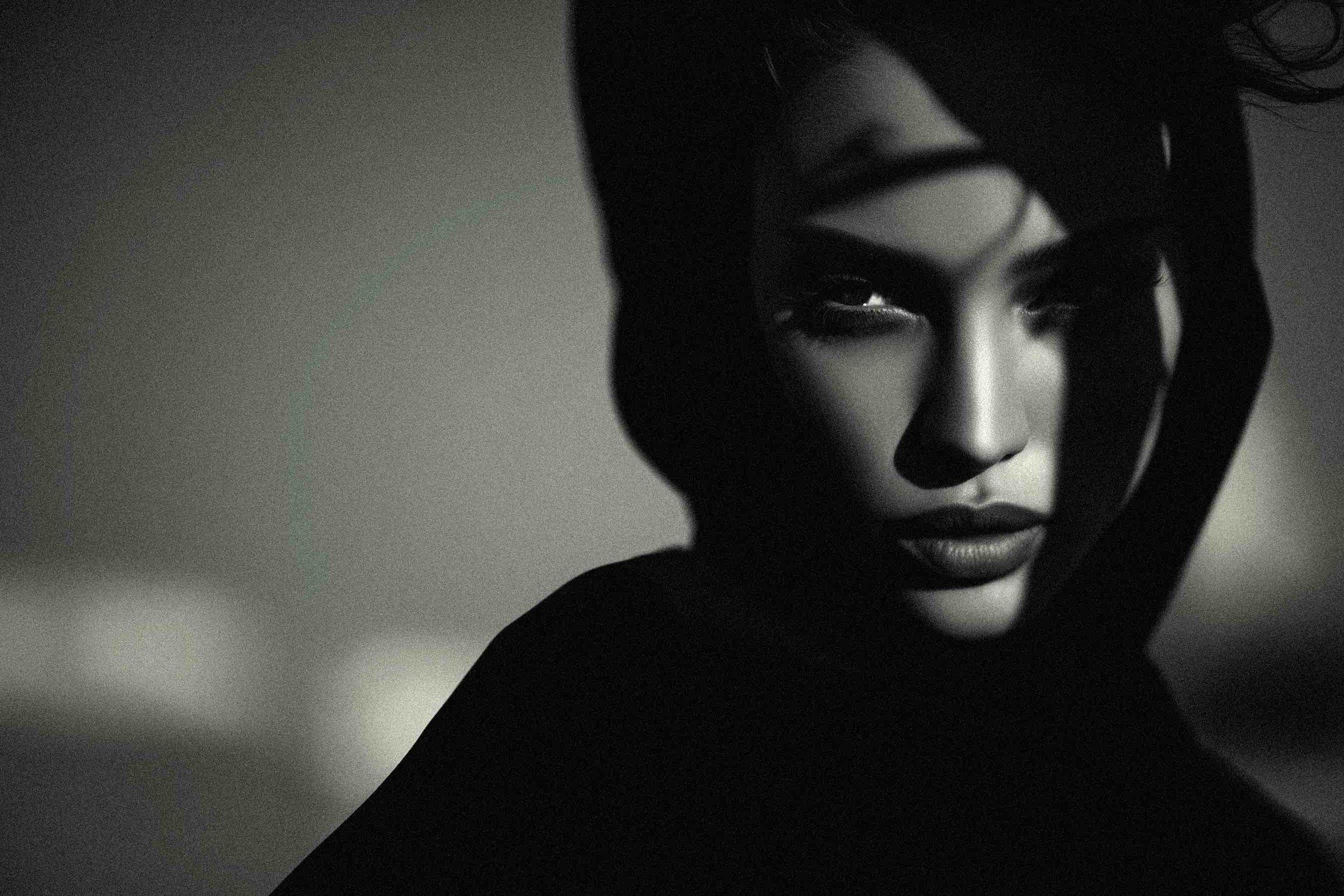 A black-and-white portrait of a woman with dark hair, intense eyes, and full lips, partially concealed behind a hood or fabric, with dramatic shadows enhancing her facial features.