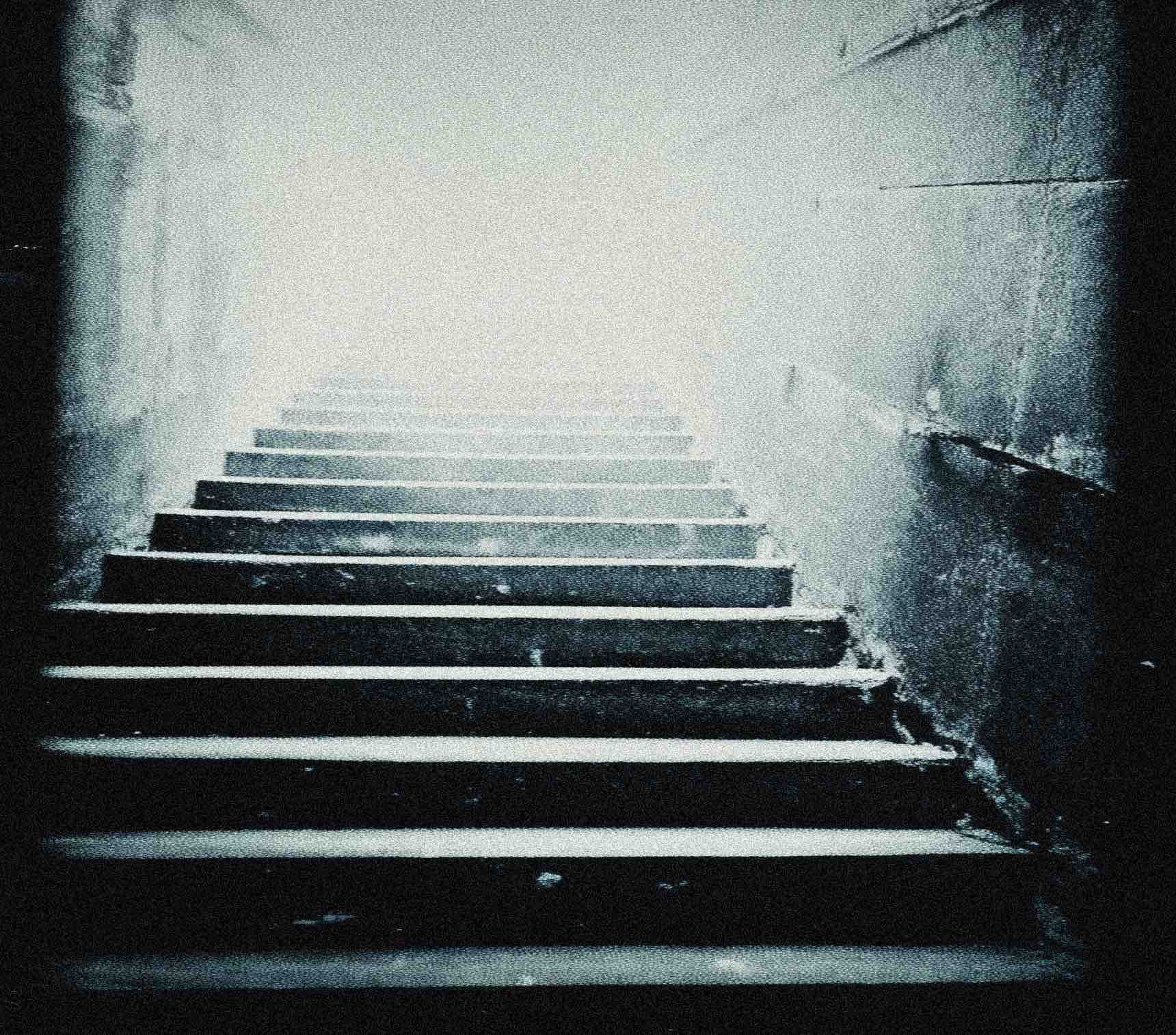 Dark staircase with black-painted steps and a white strip along the edge of each step, leading upwards into a bright, foggy area. The surrounding walls are light-colored and slightly textured.