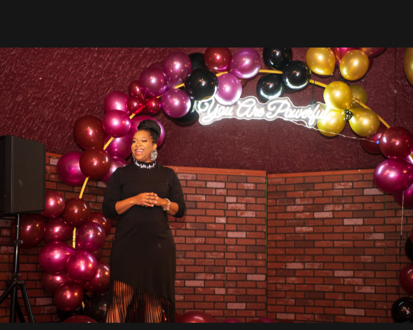 A woman in a black dress standing on a stage, smiling, with a balloon arch and a neon sign that reads 'You Are Powerful' behind her.